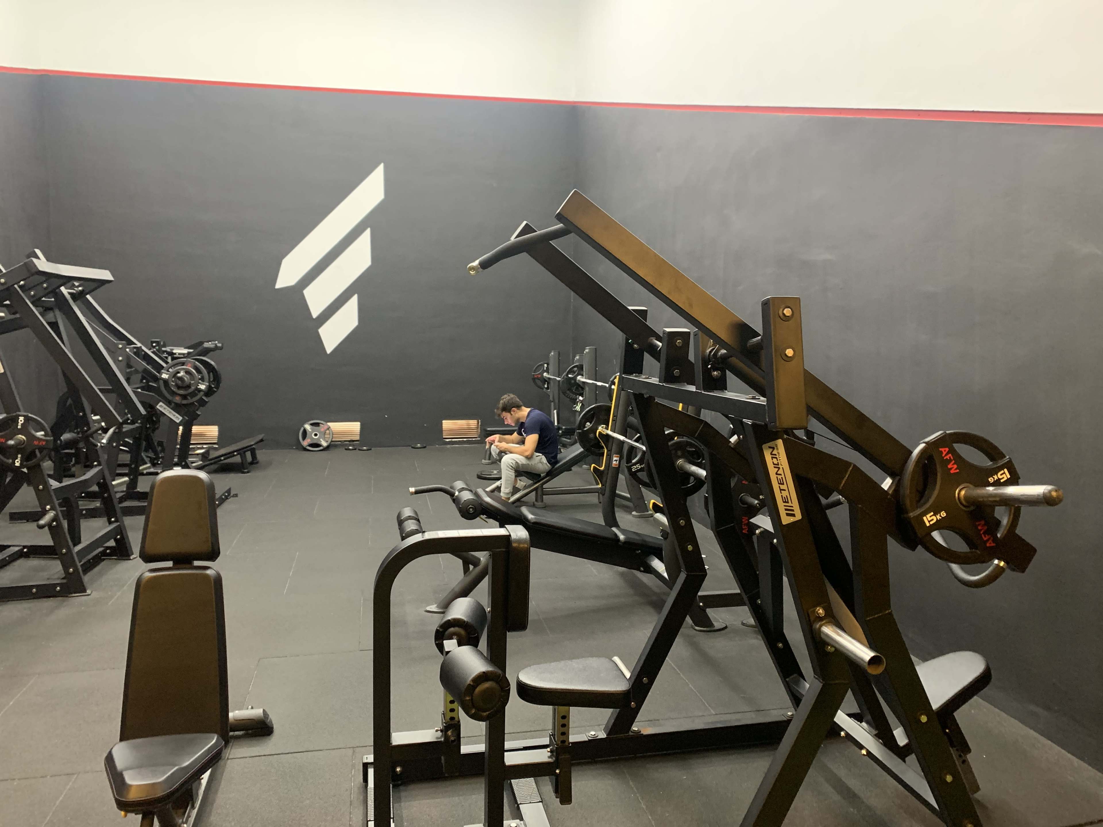A small gym space features various weightlifting machines and a person seated near a wall, focused on their phone.