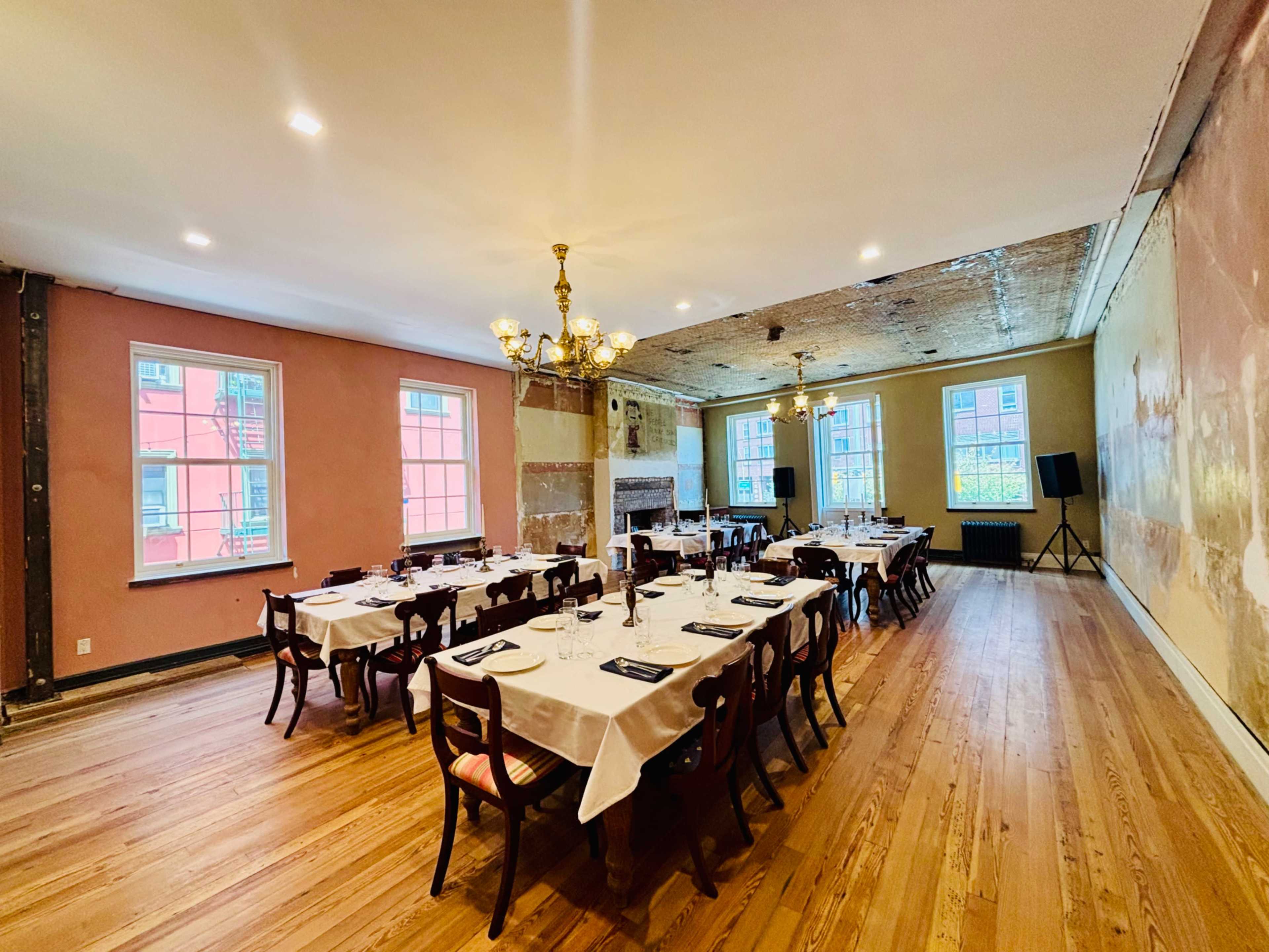 The image shows a dining room set up with multiple tables covered in white tablecloths, arranged in a rectangular formation, surrounded by exposed wooden floors and traditional decor elements.