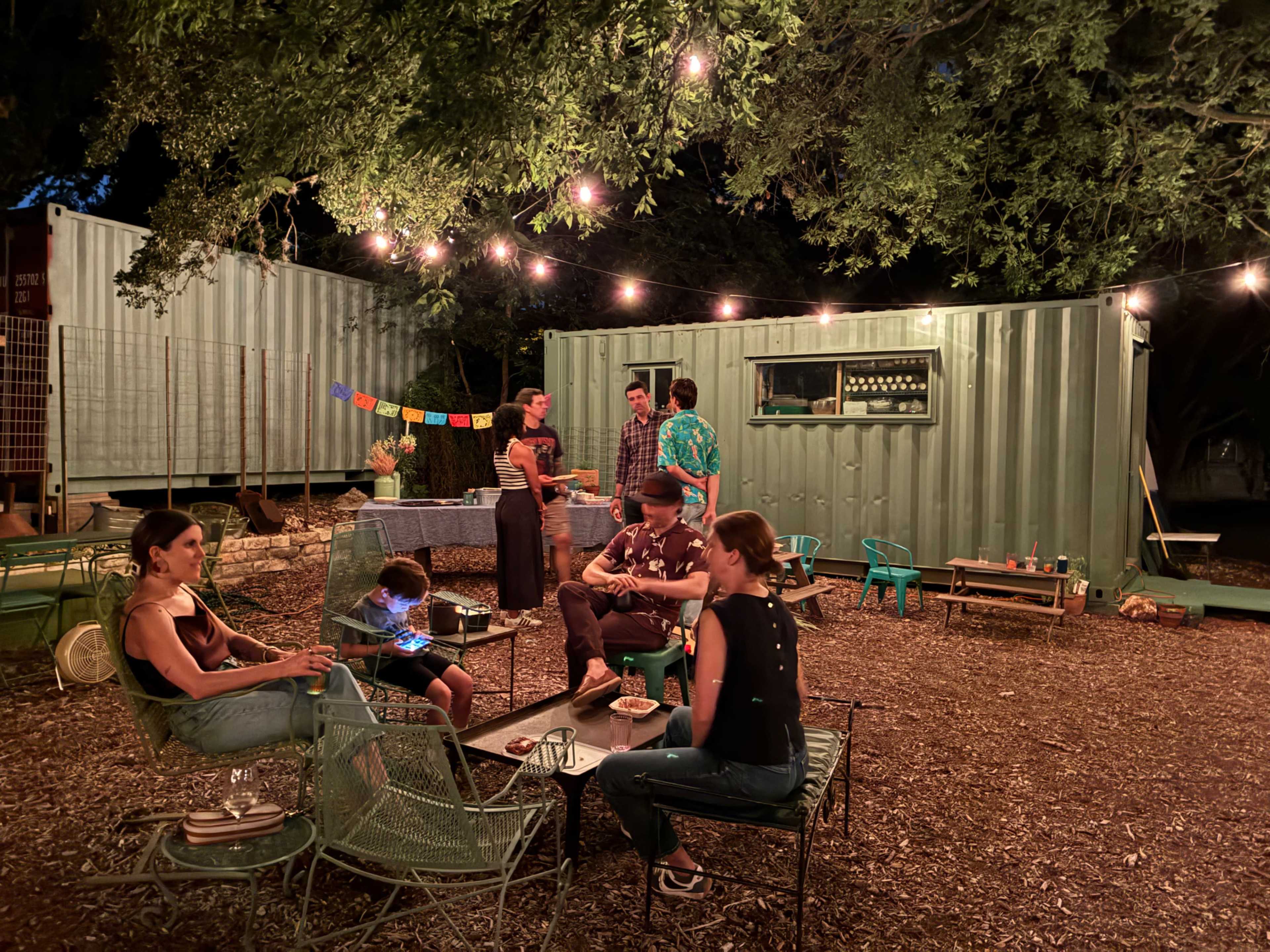 A group of people socializes in an outdoor setting illuminated by string lights, surrounded by shipping containers and seated on metal chairs.
