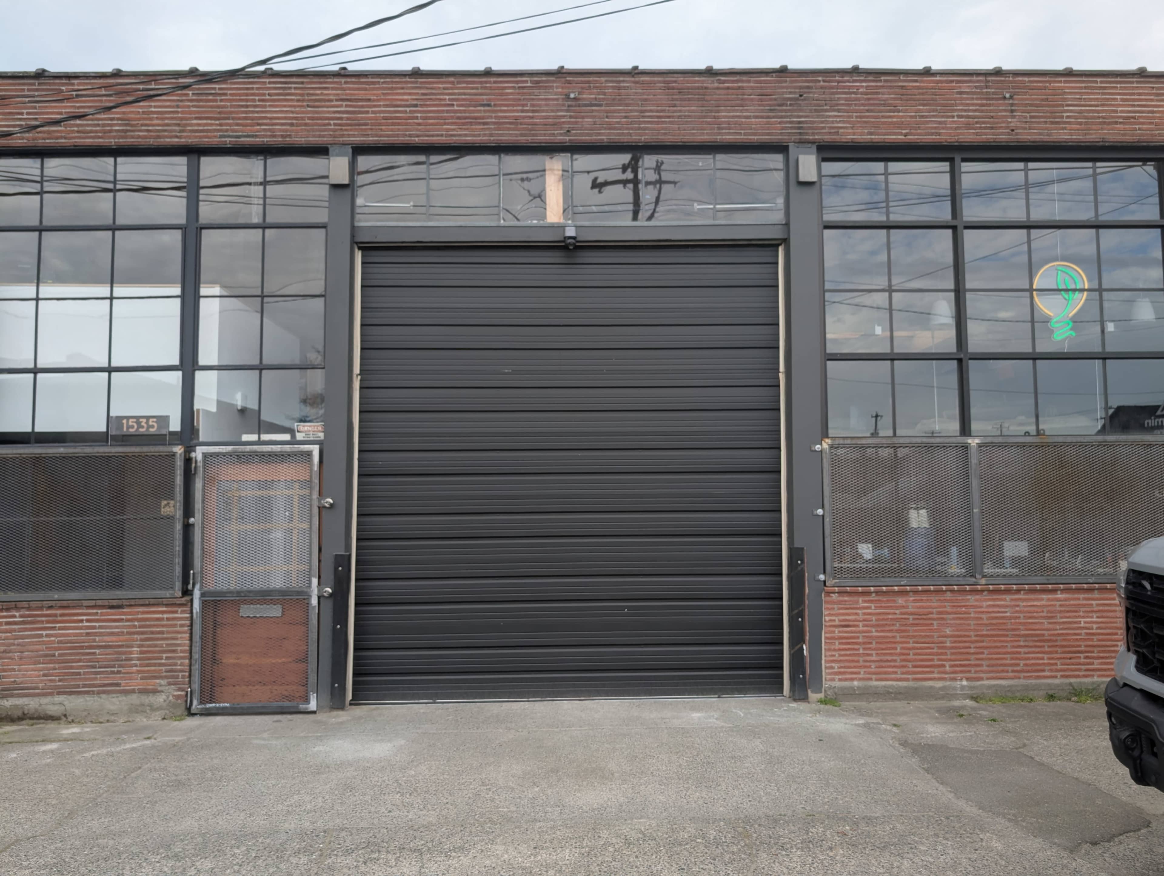 The image shows a brick industrial building with large windows and a metal roll-up door at the front.