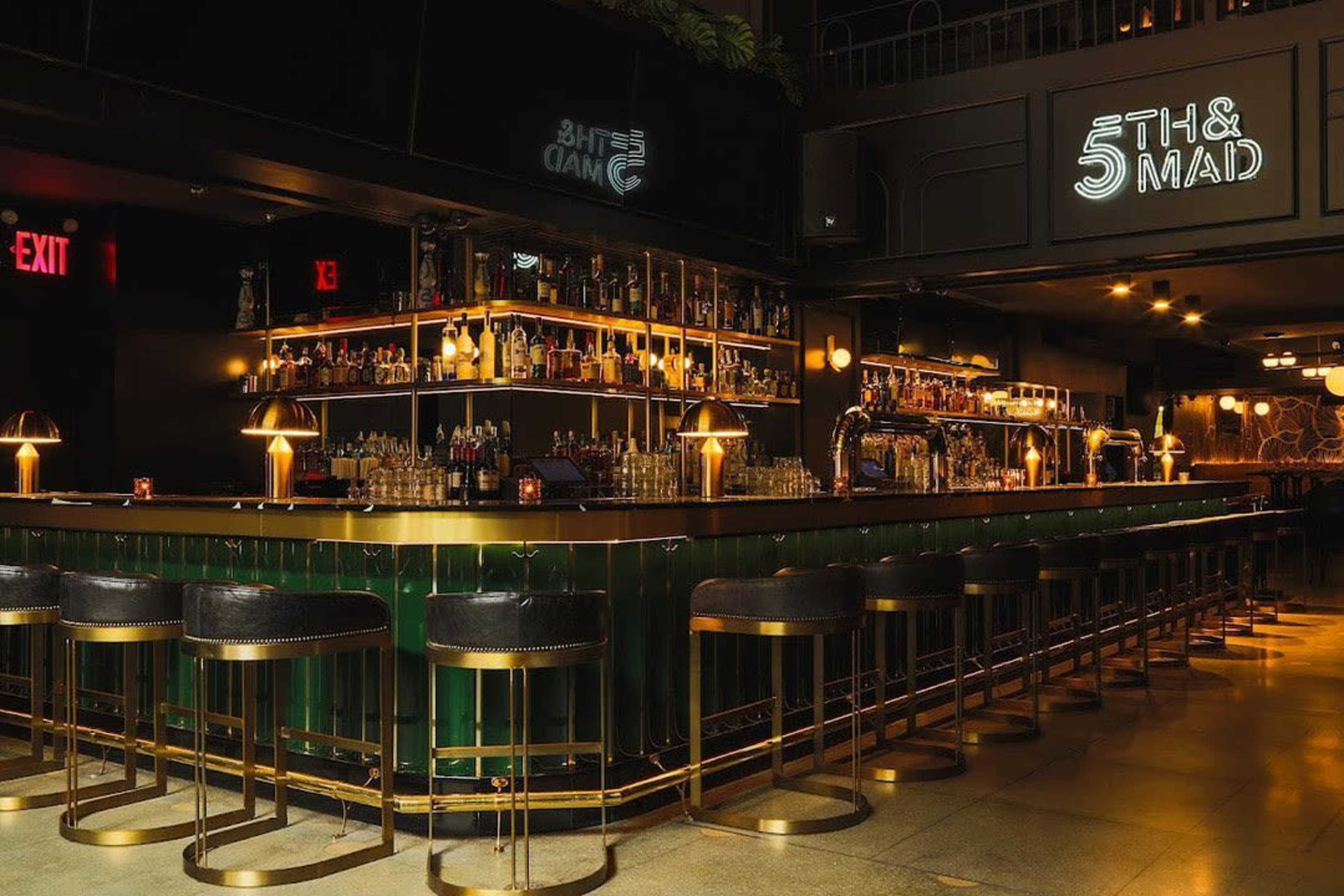 The image shows a dimly lit bar with a green and gold counter, showcasing numerous bottles on shelves behind it and stylish seating along the front.