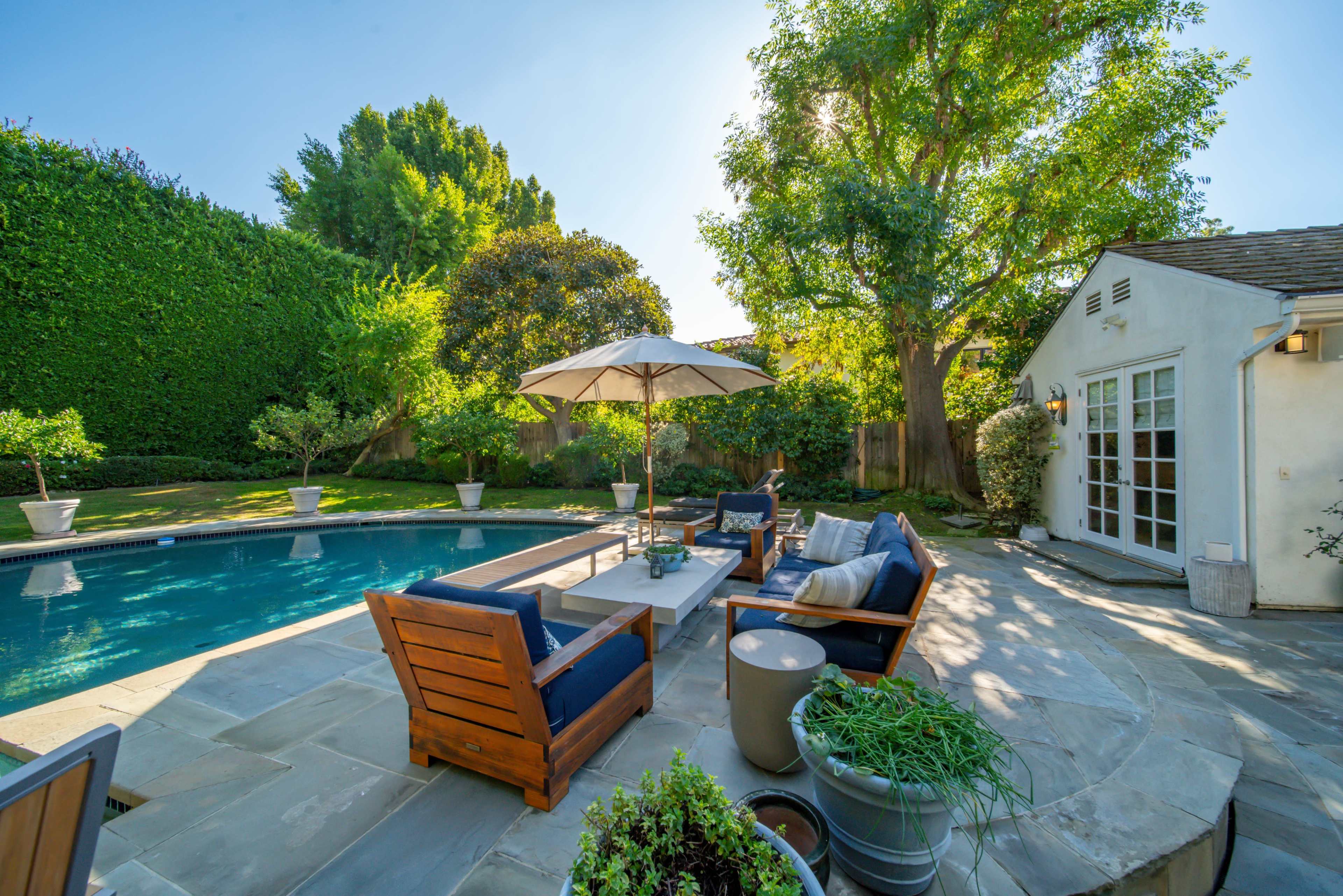 A serene backyard features a swimming pool surrounded by wooden seating, an umbrella, and lush greenery.