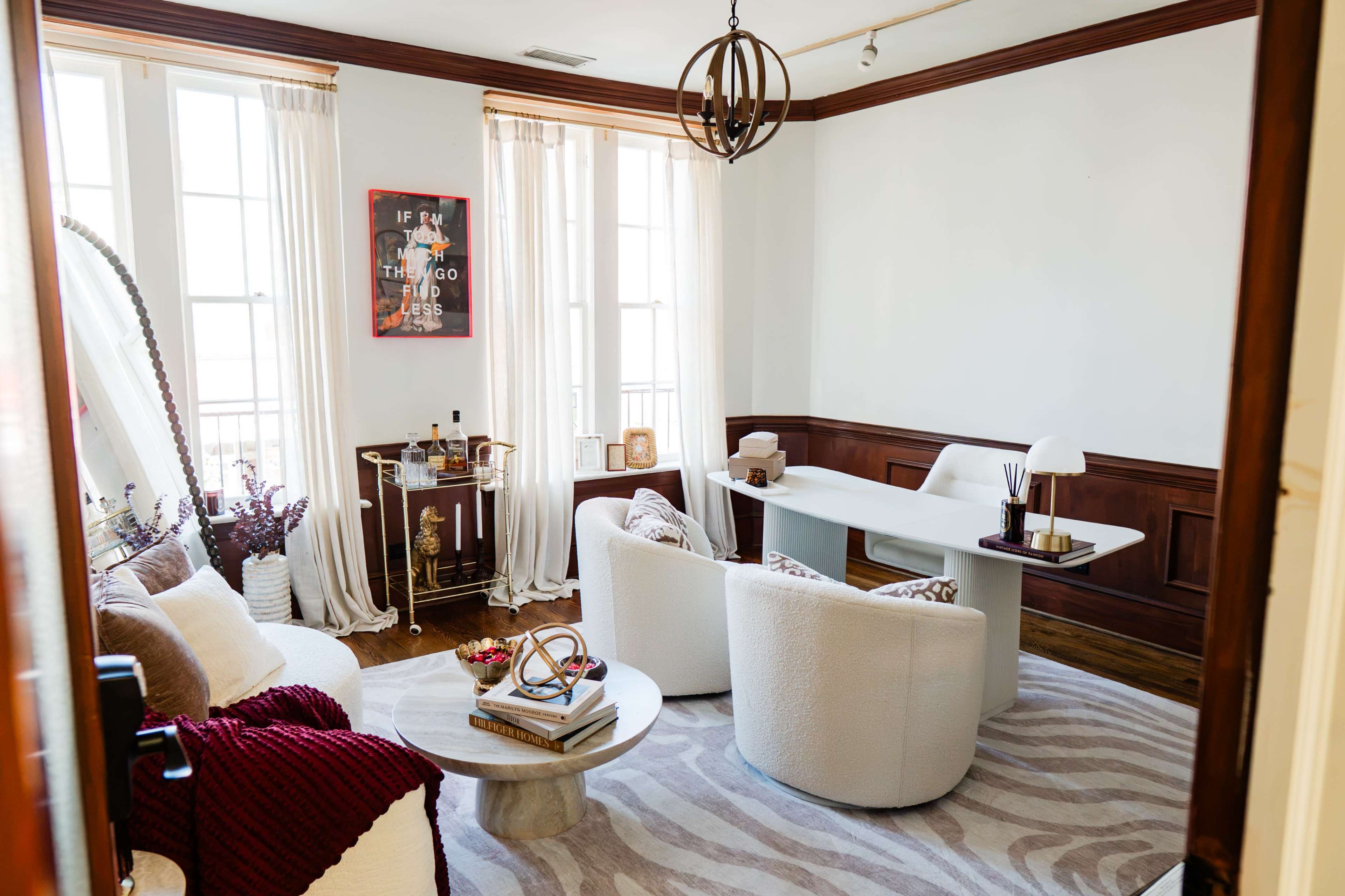 The image shows a well-lit room with large windows, featuring a modern seating area with two round sofas facing a wooden table and a desk against the wall.