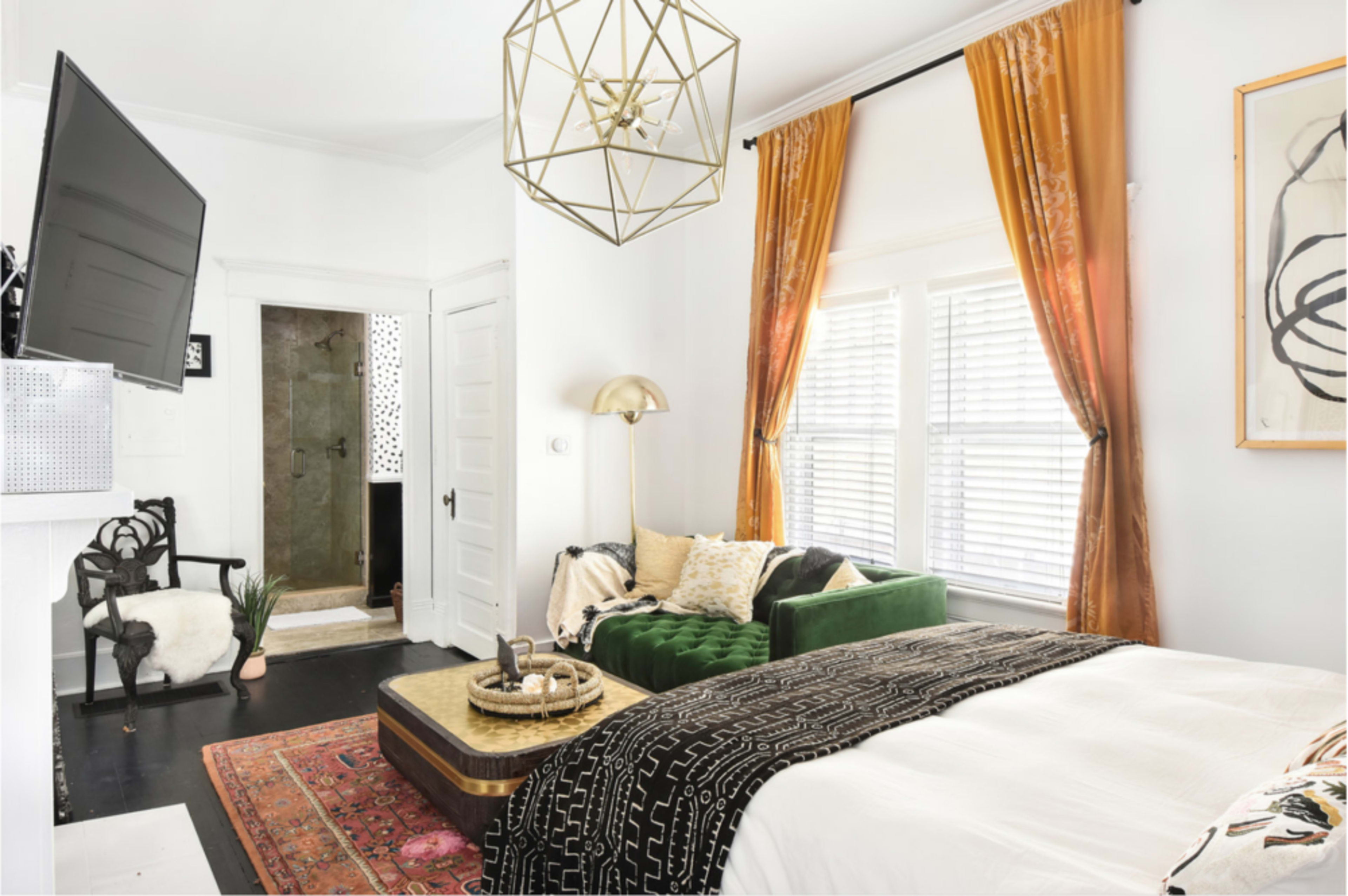 The image shows a stylish bedroom with a green velvet sofa, a large bed with patterned bedding, and decorative curtains, alongside a doorway leading to a bathroom.