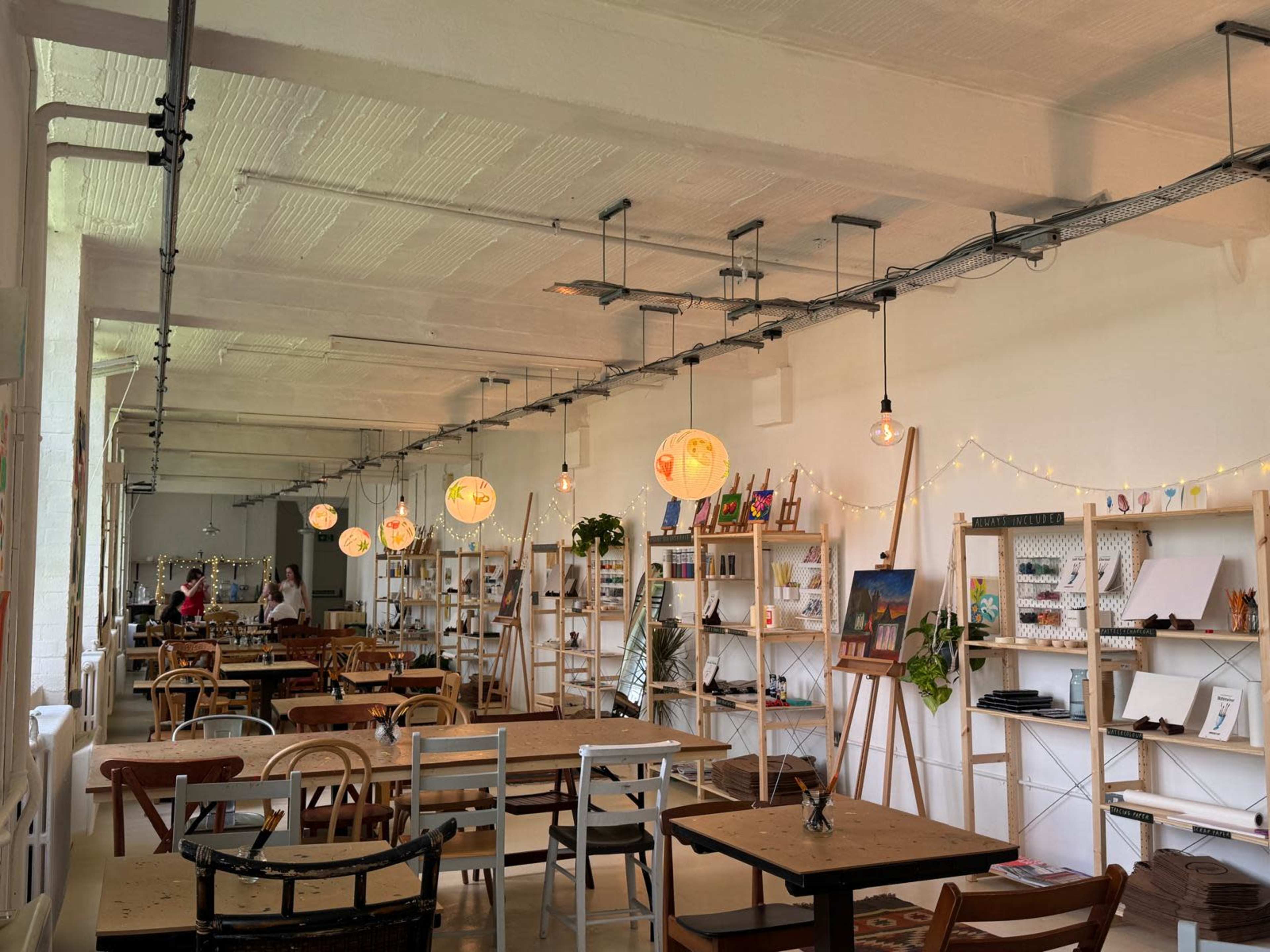 The image shows a modern café interior with wooden tables, hanging pendant lights, and decorative shelves displaying various items along the walls.