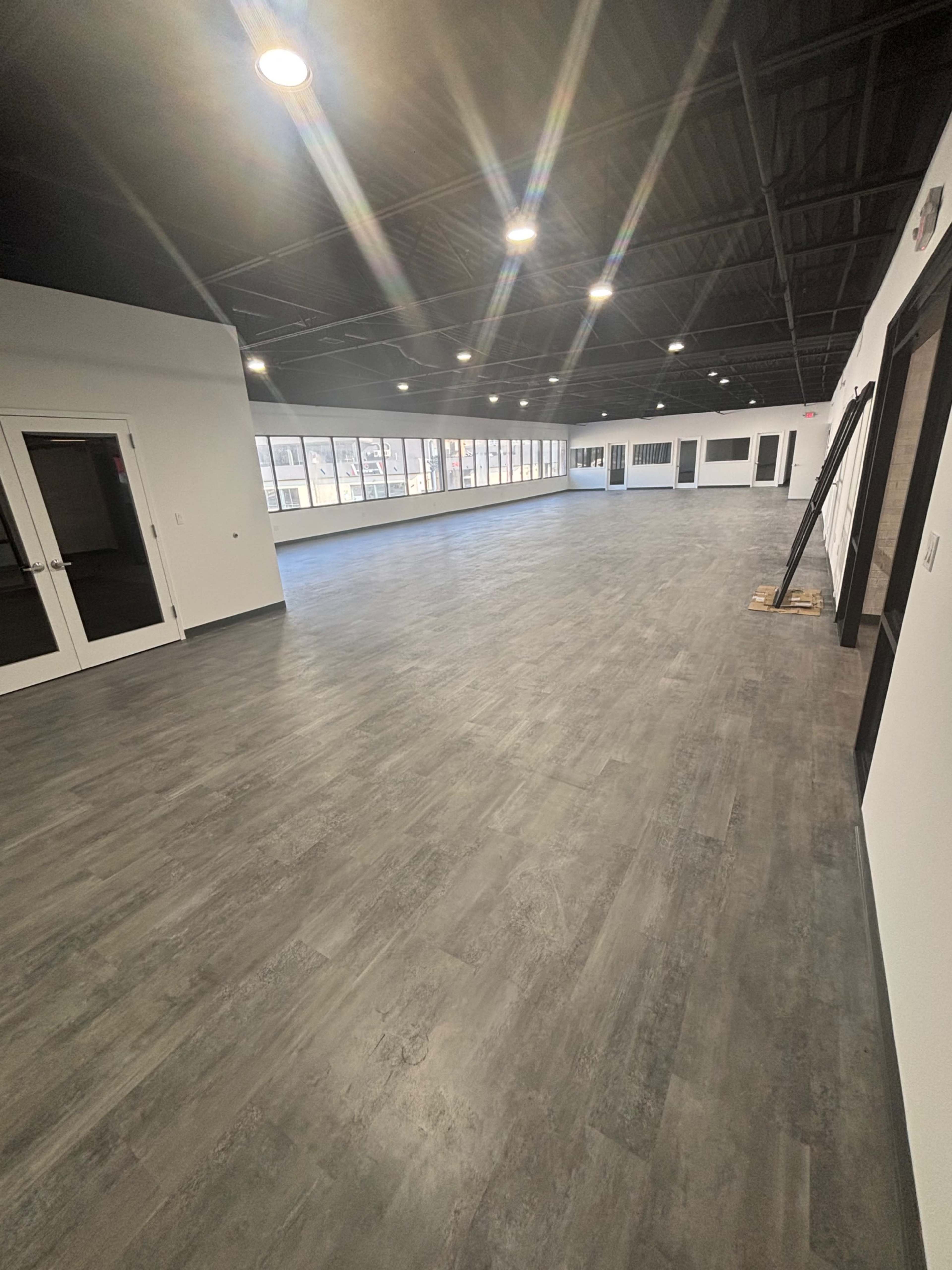 The image shows a spacious, empty room with large windows and a smooth floor, illuminated by overhead lights.