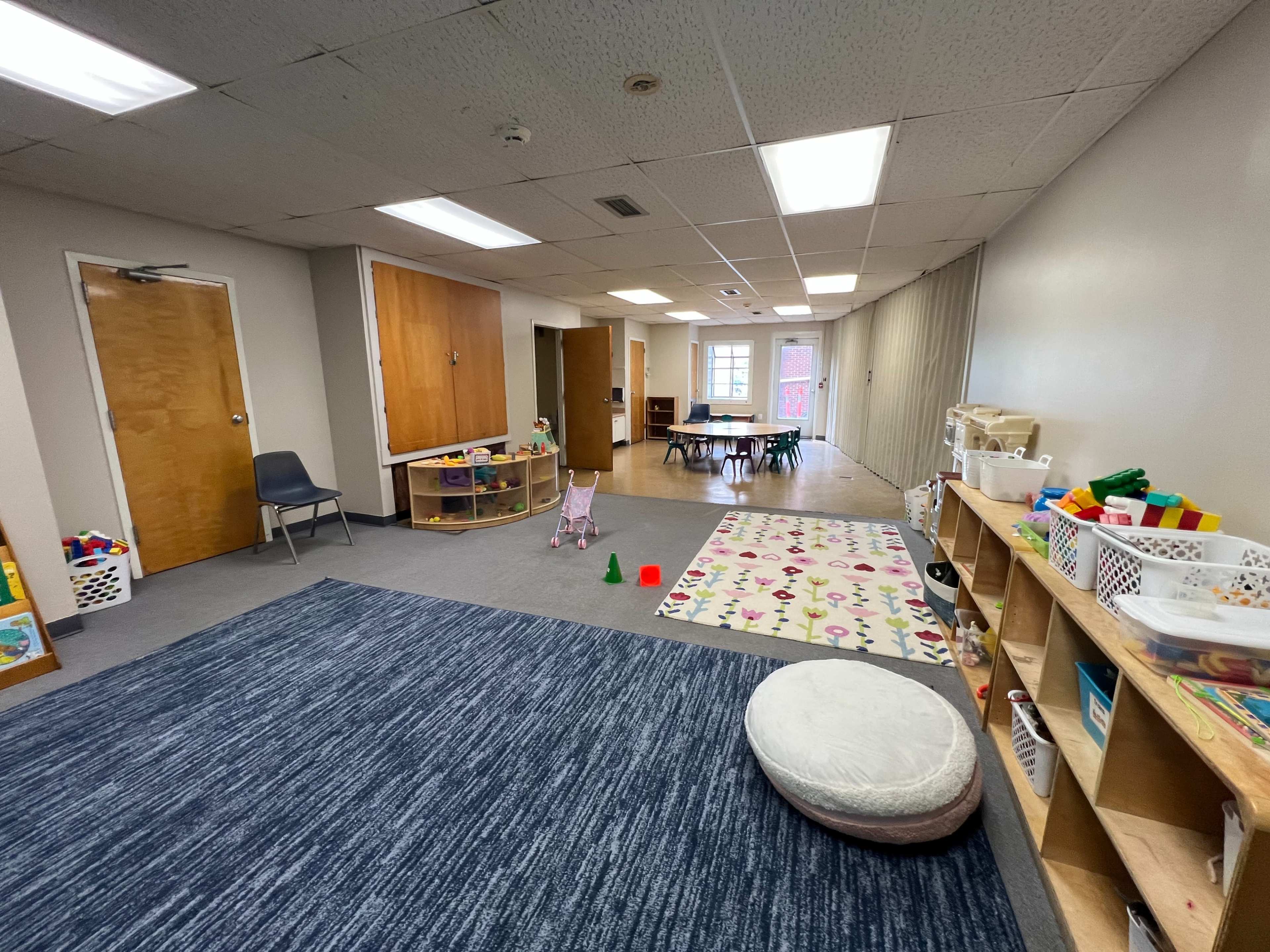 The image shows a bright classroom with play areas, furniture for children, and shelves filled with toys.