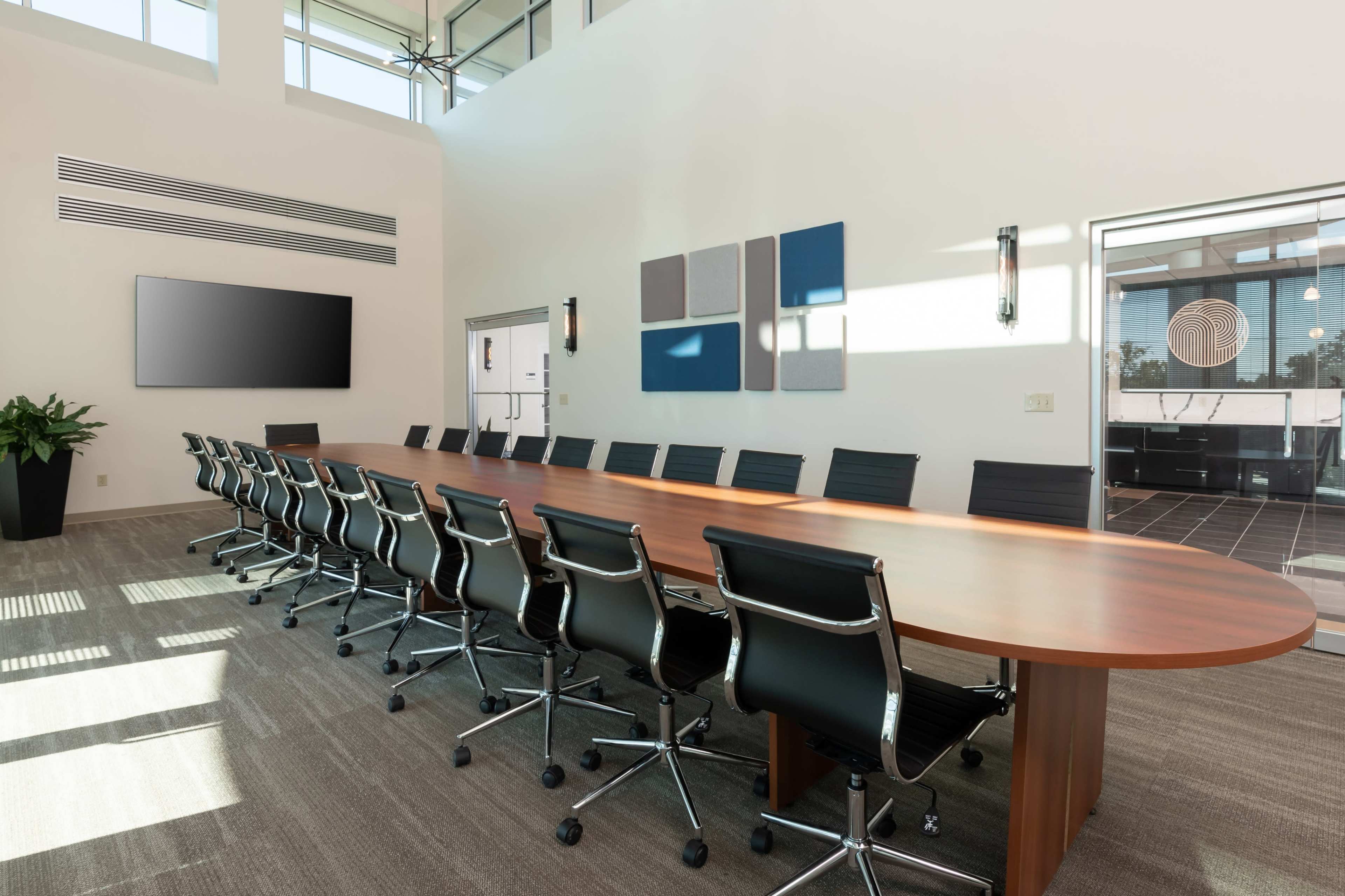 A large, modern conference room features a long wooden table with black rolling chairs and a wall-mounted screen.