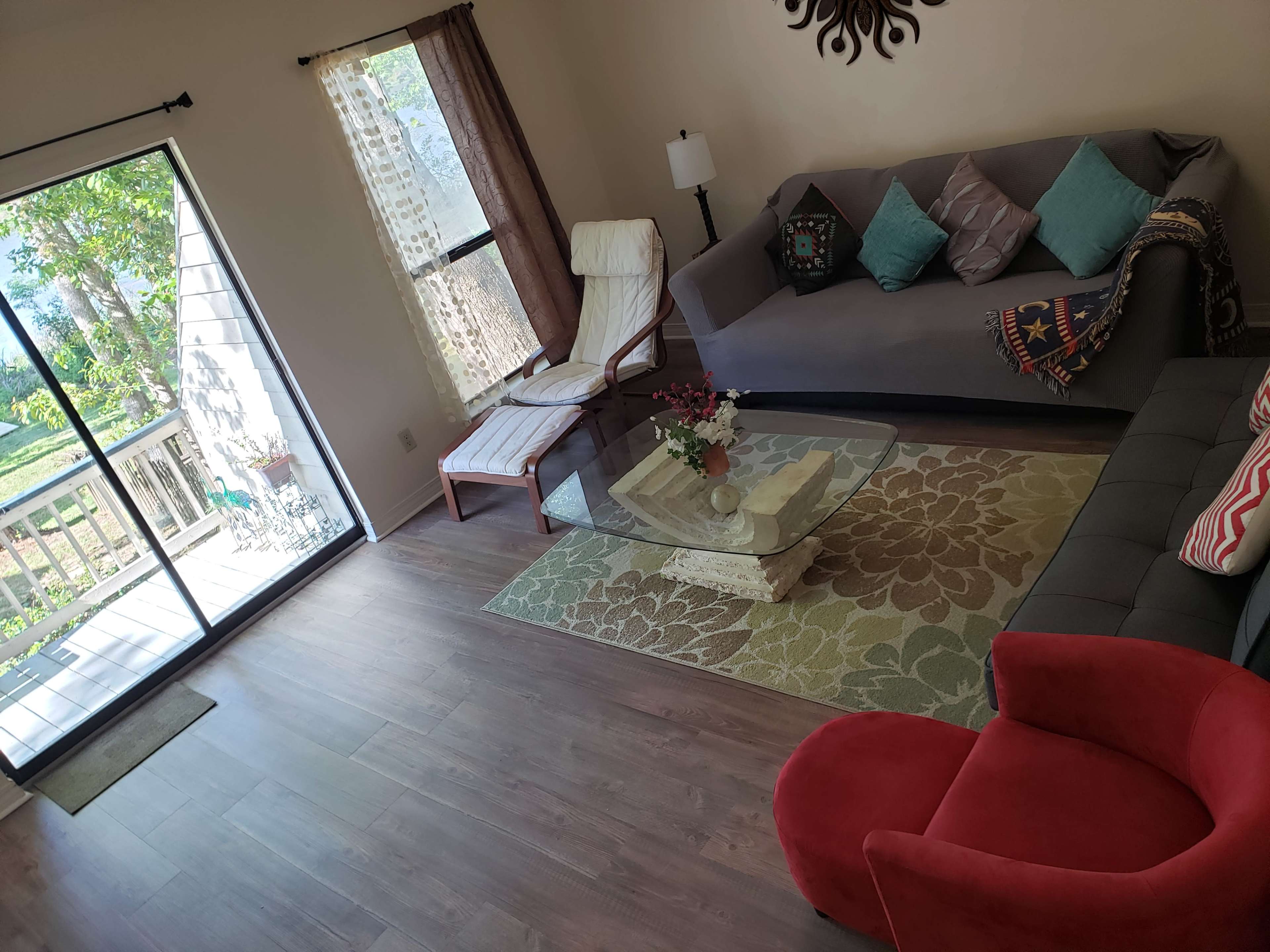 The image shows a living room with a glass coffee table, a gray sofa, a red armchair, and large windows letting in natural light.