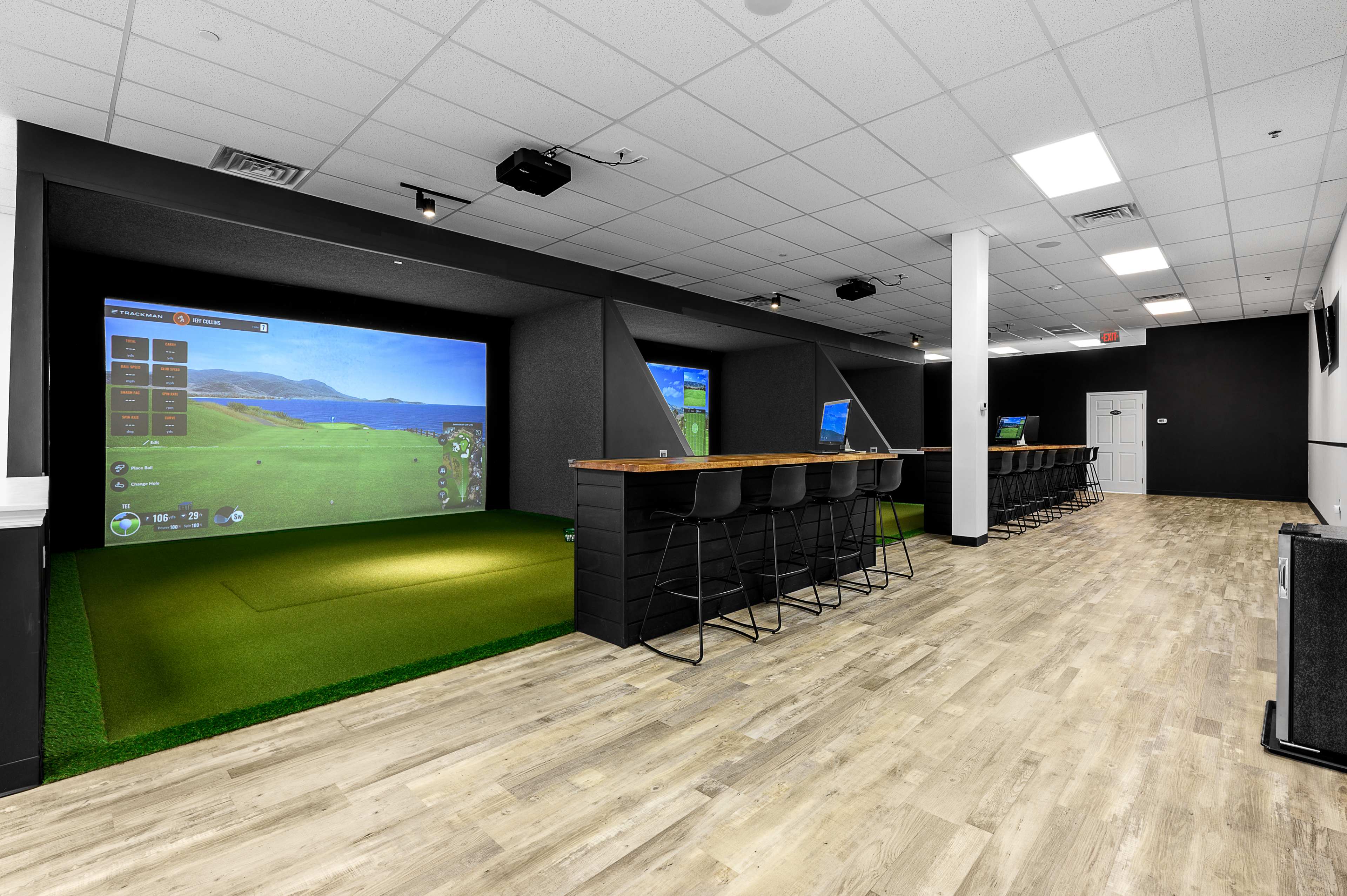 The image shows an indoor golf simulation room featuring two large screens displaying golf courses and a lounge area with seating and a counter.