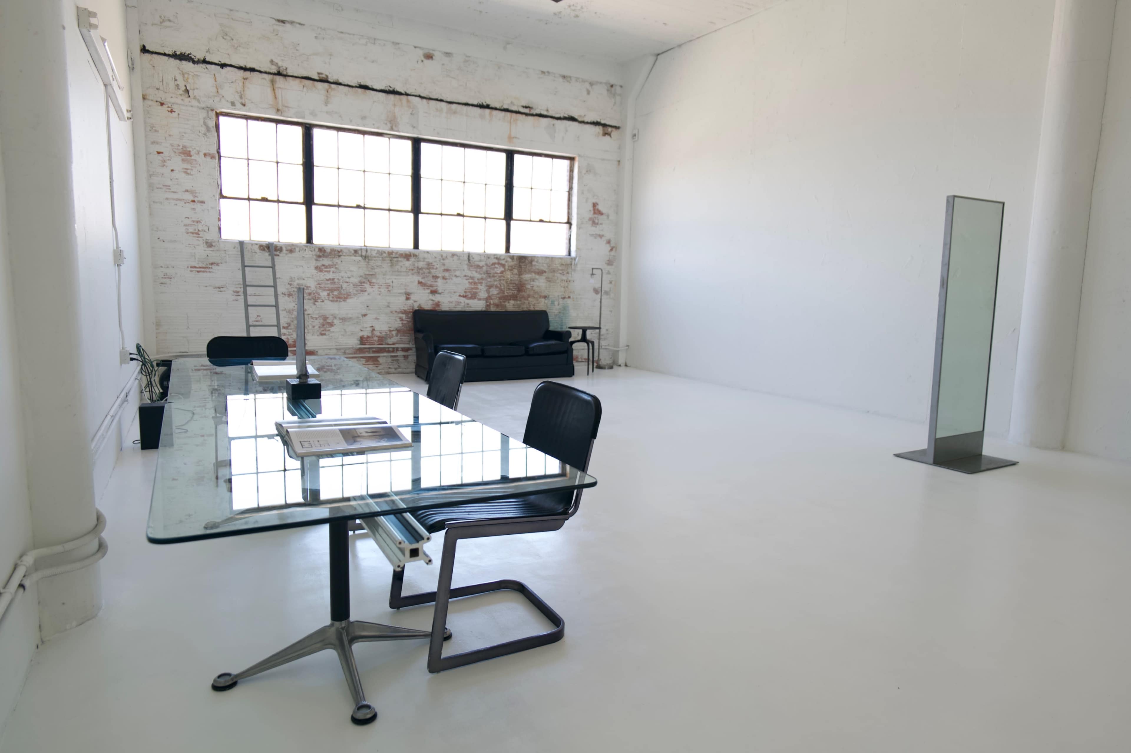 The image shows a minimalist office space with a glass table, black chairs, a sofa, and large windows along a white brick wall.