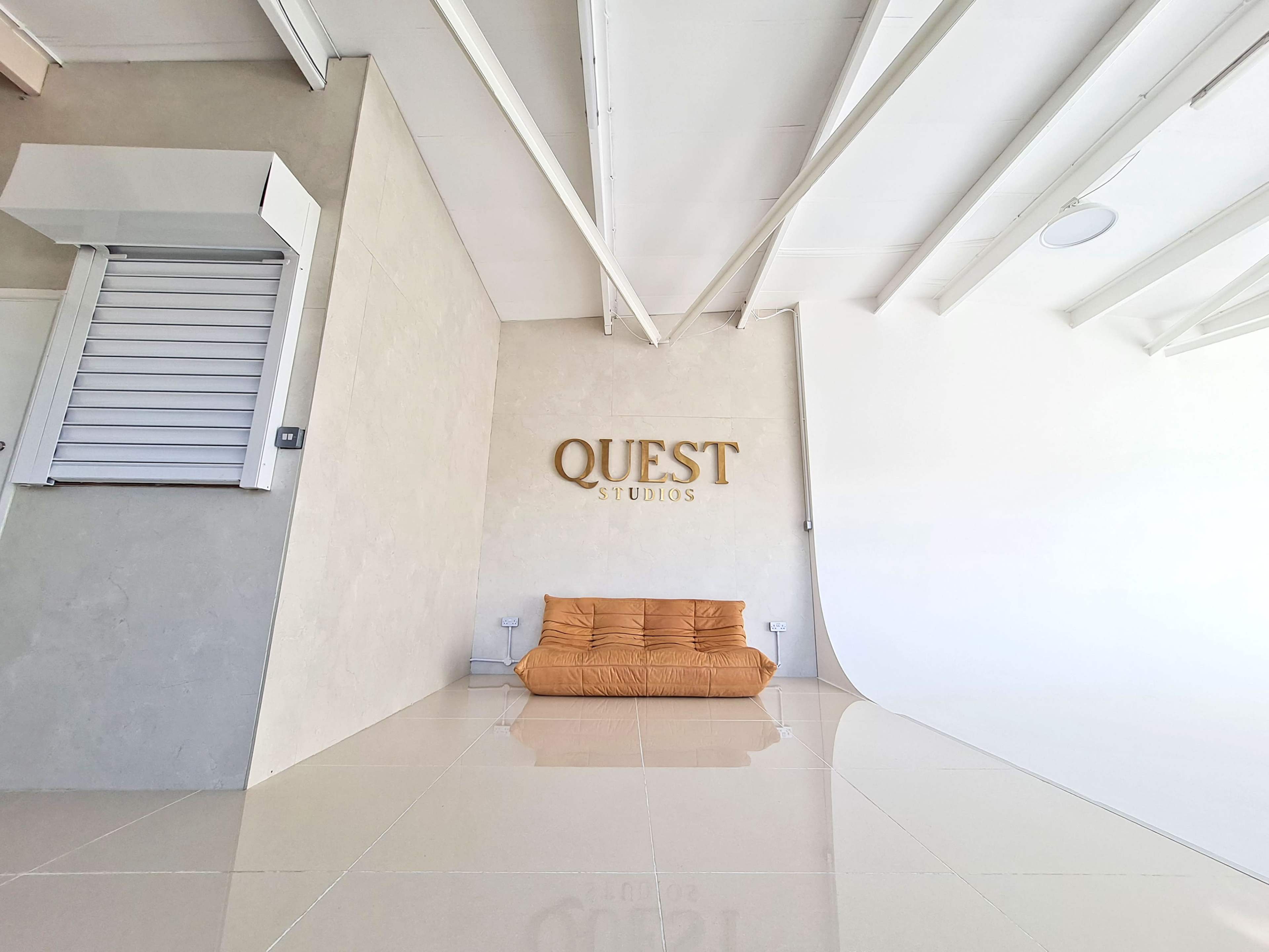 A minimalist studio space with a large white backdrop and a brown sofa against a light-colored wall featuring the word "QUEST" in gold letters.