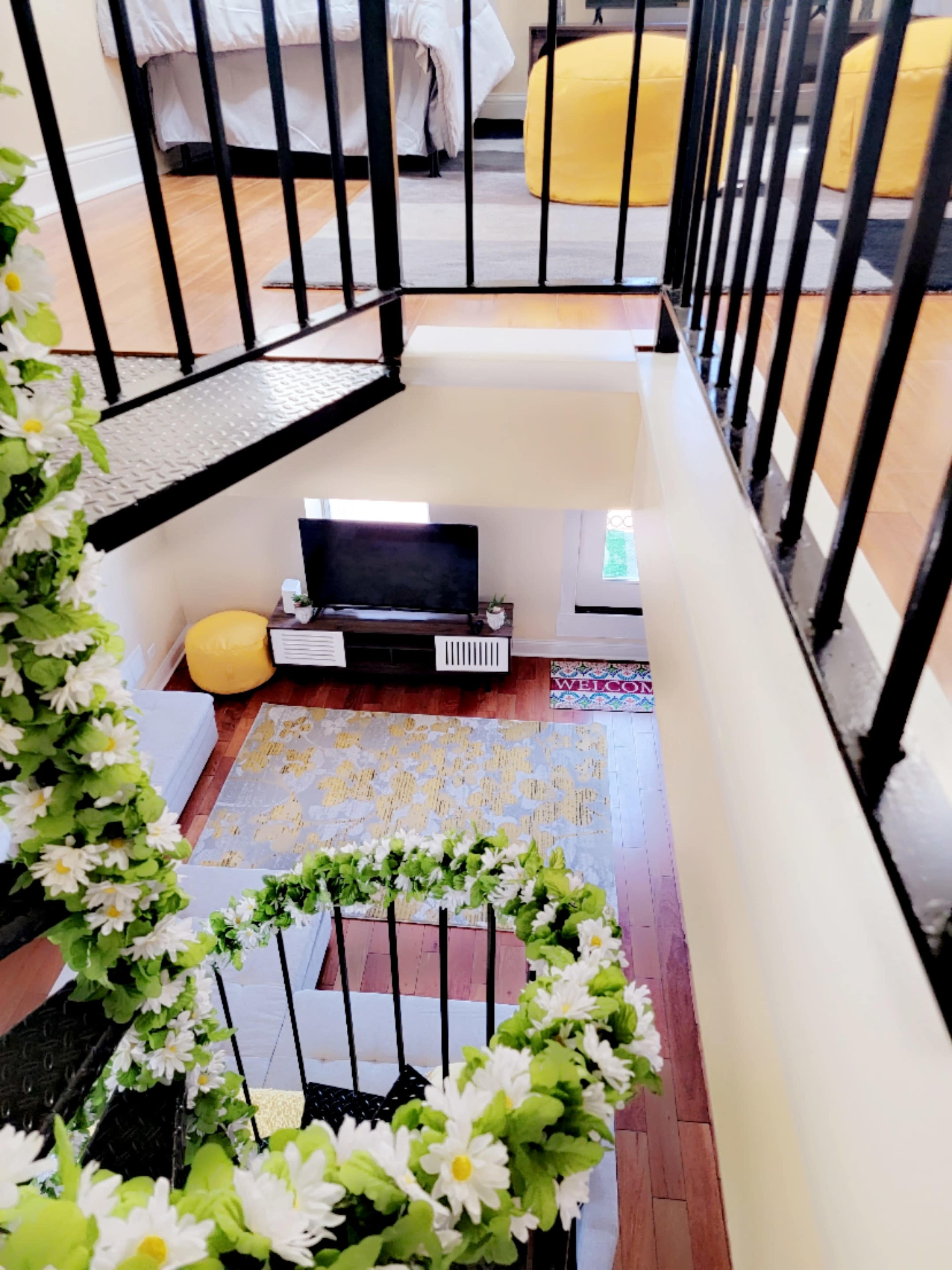 A spiral staircase adorned with greenery leads to a living space featuring a television, a yellow ottoman, and a patterned rug.