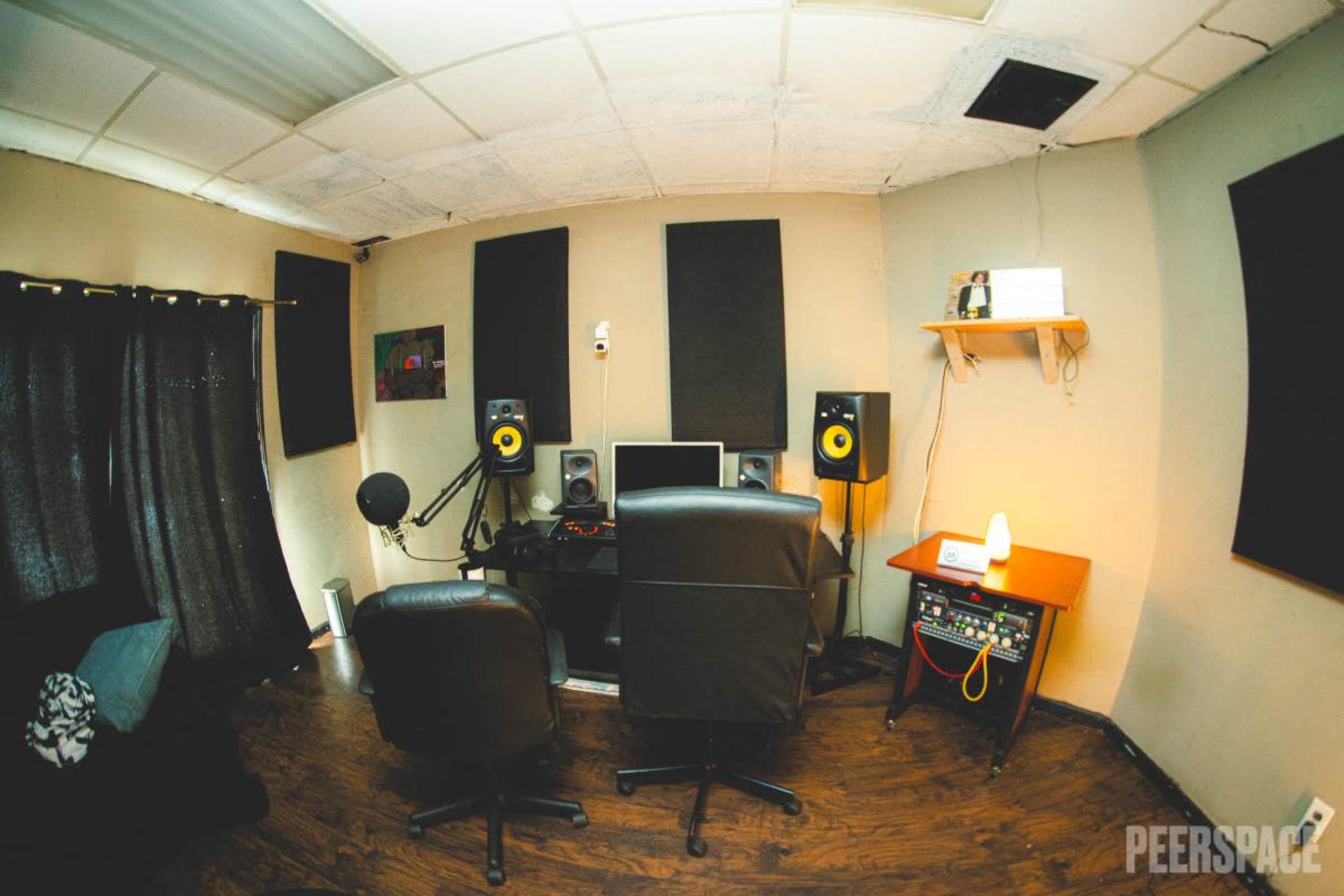 The image shows a small, organized recording studio with two monitors, audio speakers, and chairs arranged around a central desk.