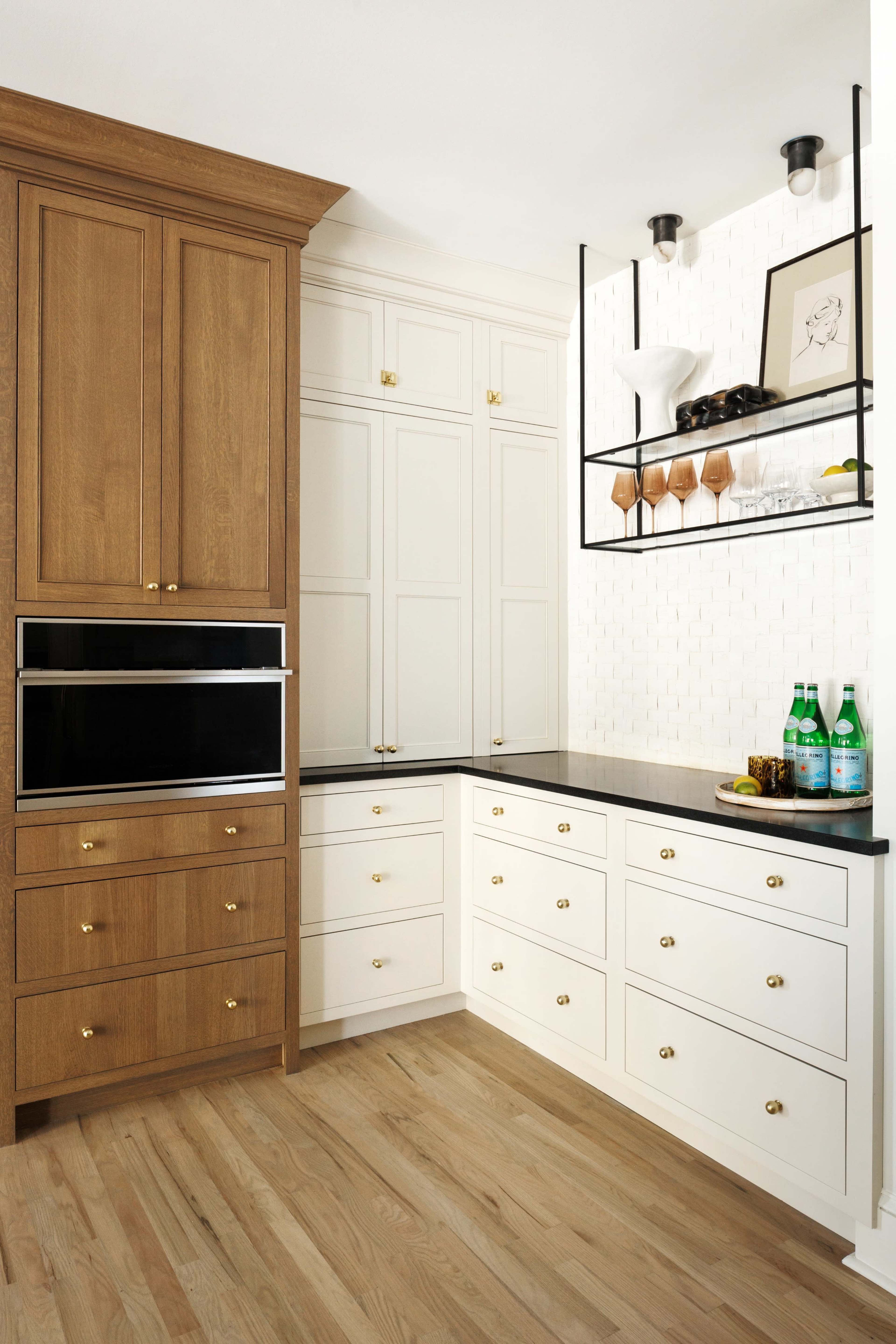 The image shows a modern kitchen featuring a combination of wooden and white cabinetry, a black countertop, and a wall shelf displaying glassware and beverages.