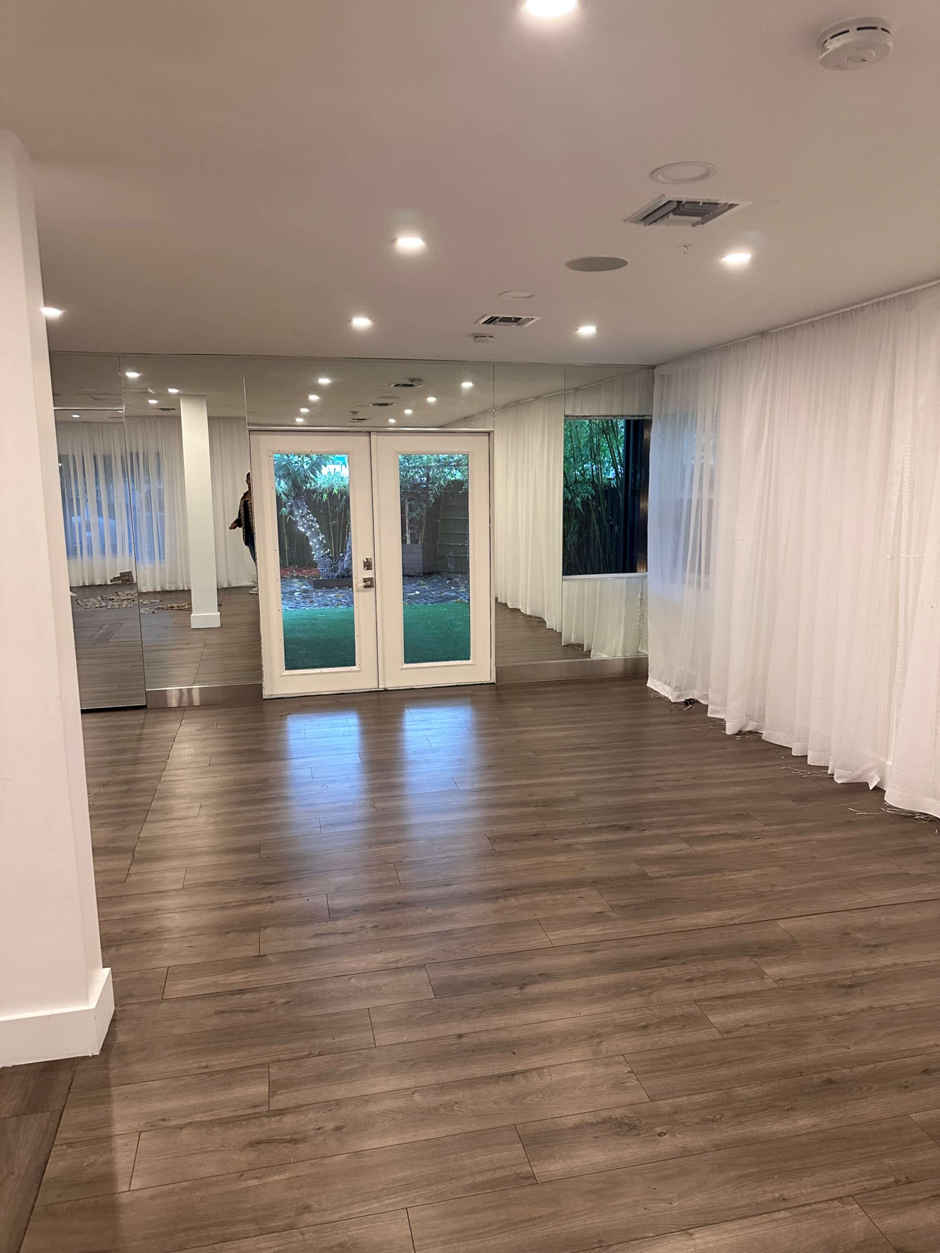 A spacious interior room with wooden flooring, large mirrors on one wall, and sliding glass doors leading to a garden area, all illuminated by ceiling lights.