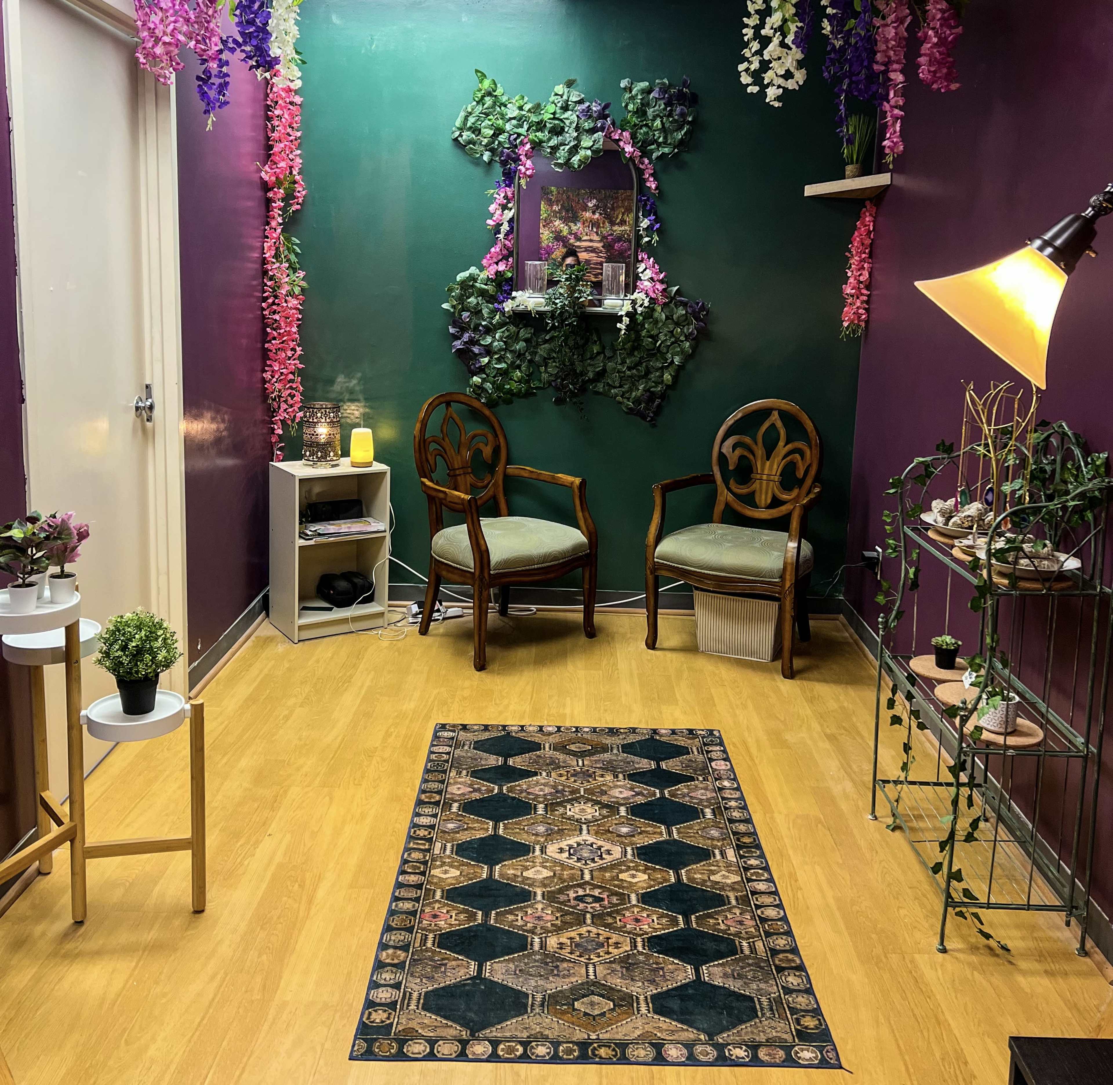 The image shows a cozy waiting area with two wooden chairs, green and purple walls adorned with faux plants, and a decorative rug on a wooden floor.