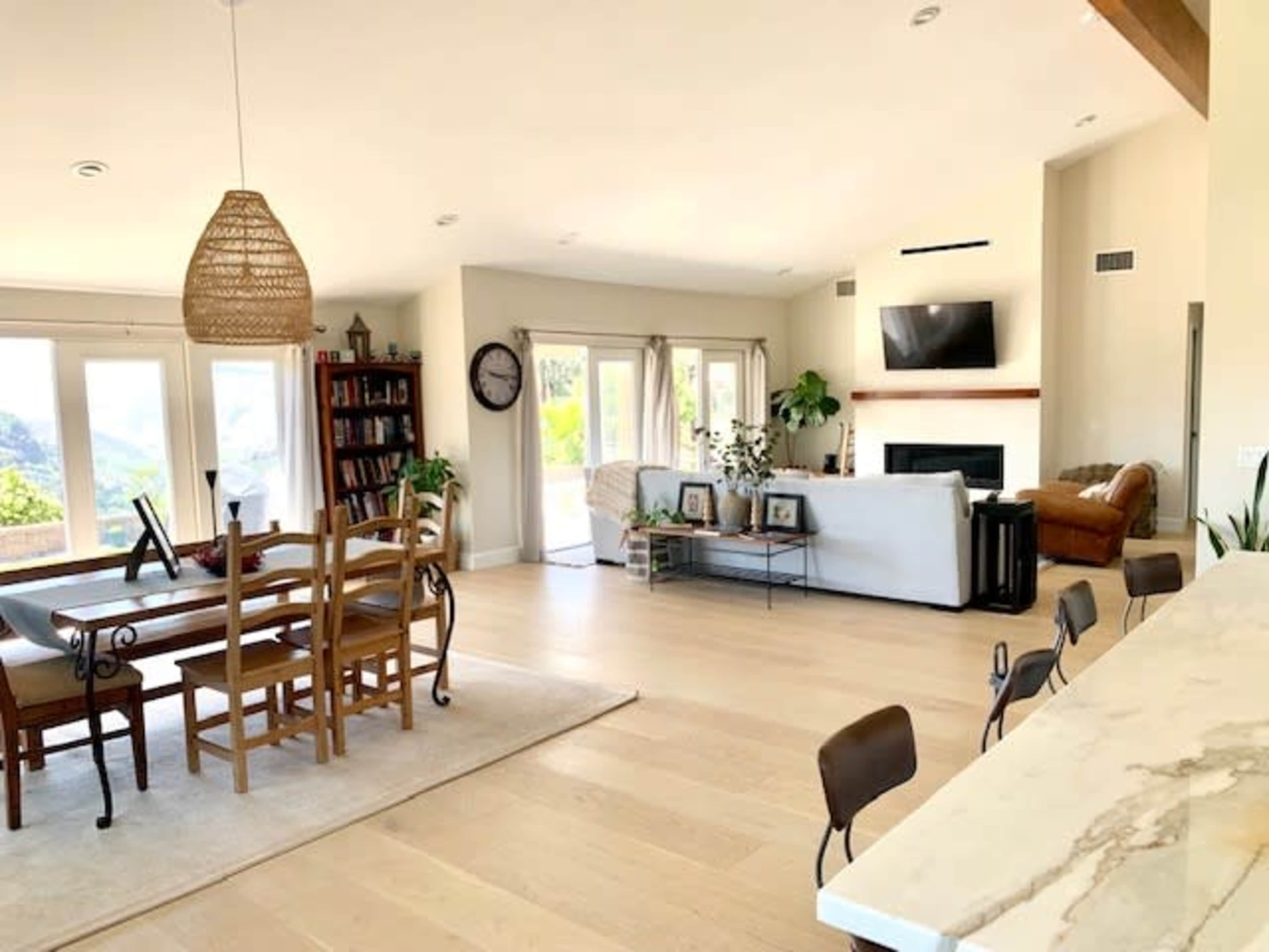 A modern open-concept living space with a dining area, a cozy sitting area featuring a fireplace, and large windows overlooking a scenic view, all with light-colored wood flooring.