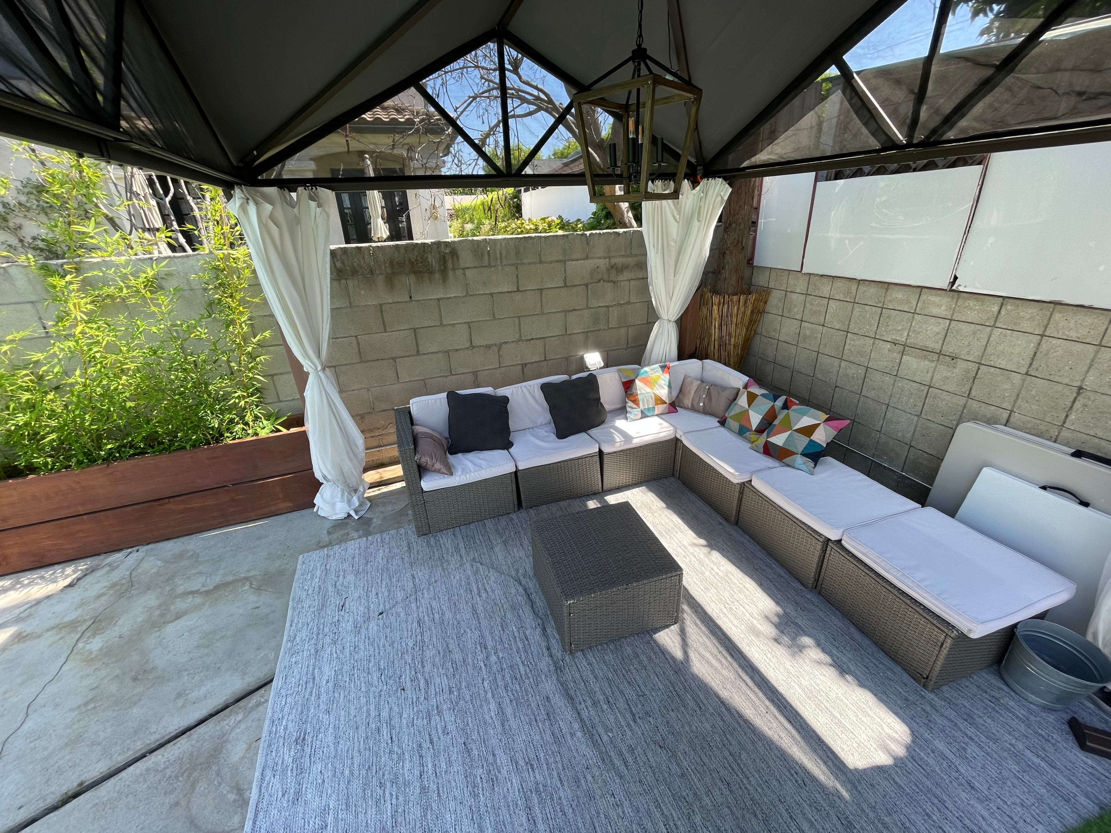 A shaded outdoor seating area with a sectional couch, a coffee table, and light curtains near a concrete wall and greenery.