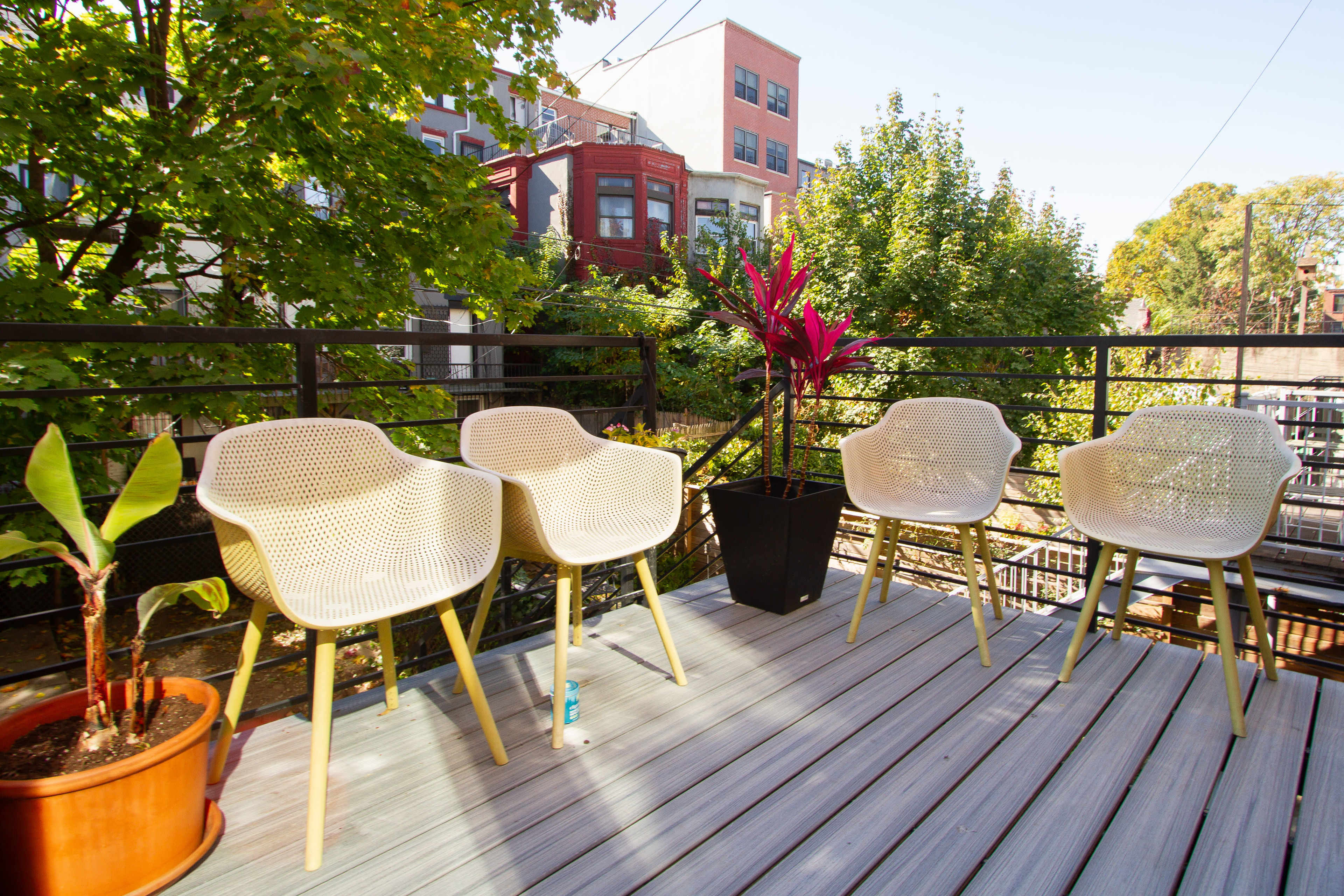 A deck features four white chairs arranged around a potted plant in a vibrant green setting with nearby buildings.