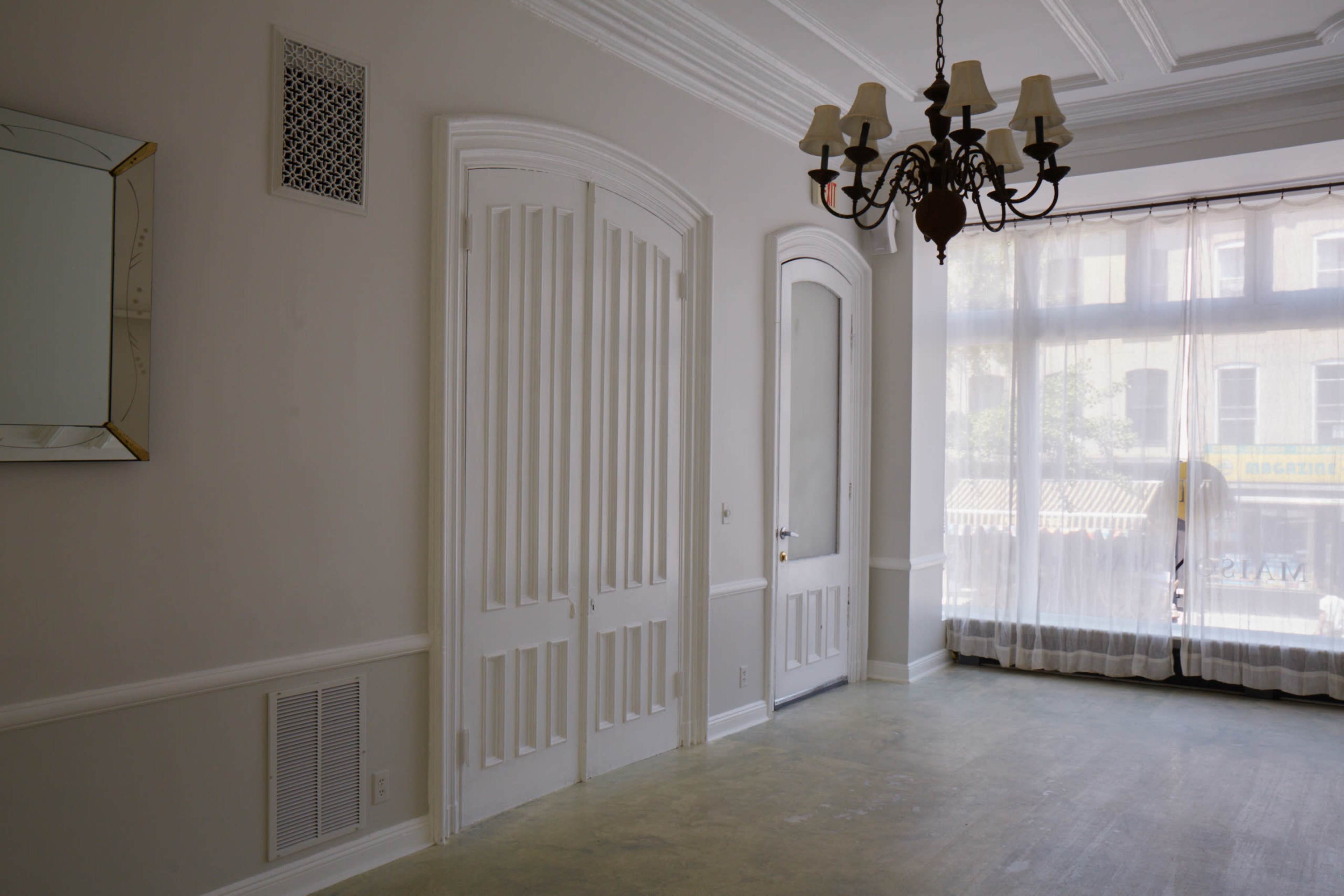 The image shows a spacious room with white walls, a large window covered by sheer curtains, two doors, a chandelier, and a decorative mirror on the wall.