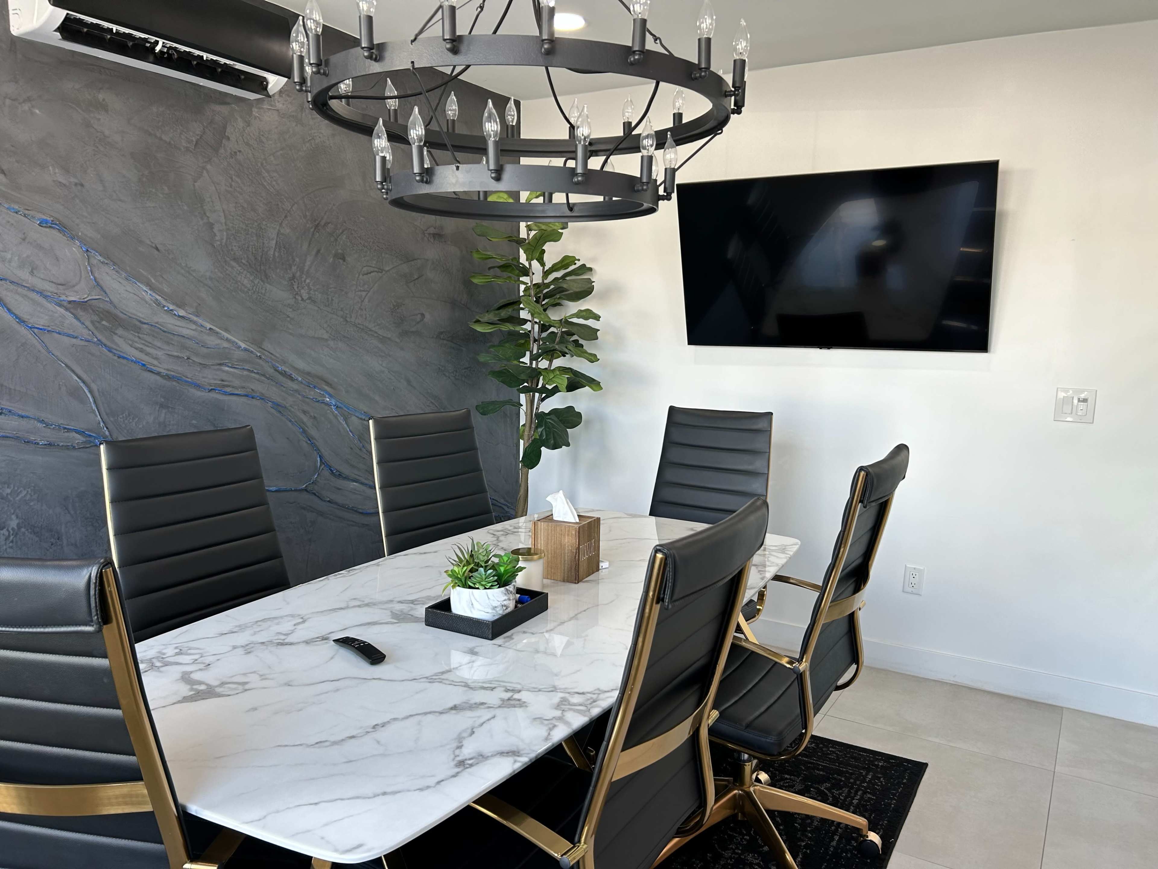 A modern conference room with a marble table, six black chairs, a large circular chandelier, a plant, and a wall-mounted TV.