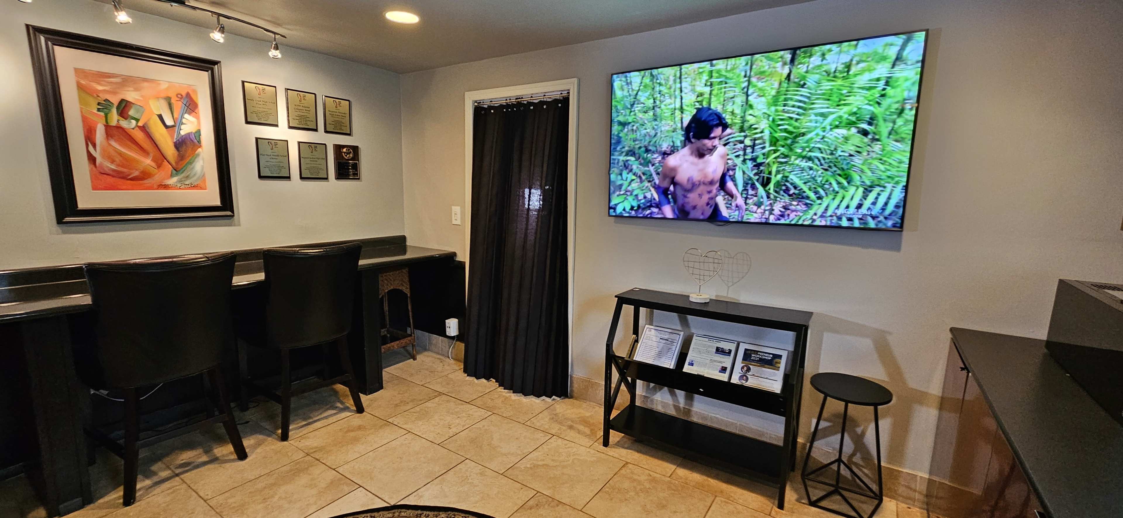 A modern interior with a wall-mounted TV displaying a nature scene, a small black table, and a bar-style seating area with high-back chairs, alongside a wall adorned with framed plaques and artwork.