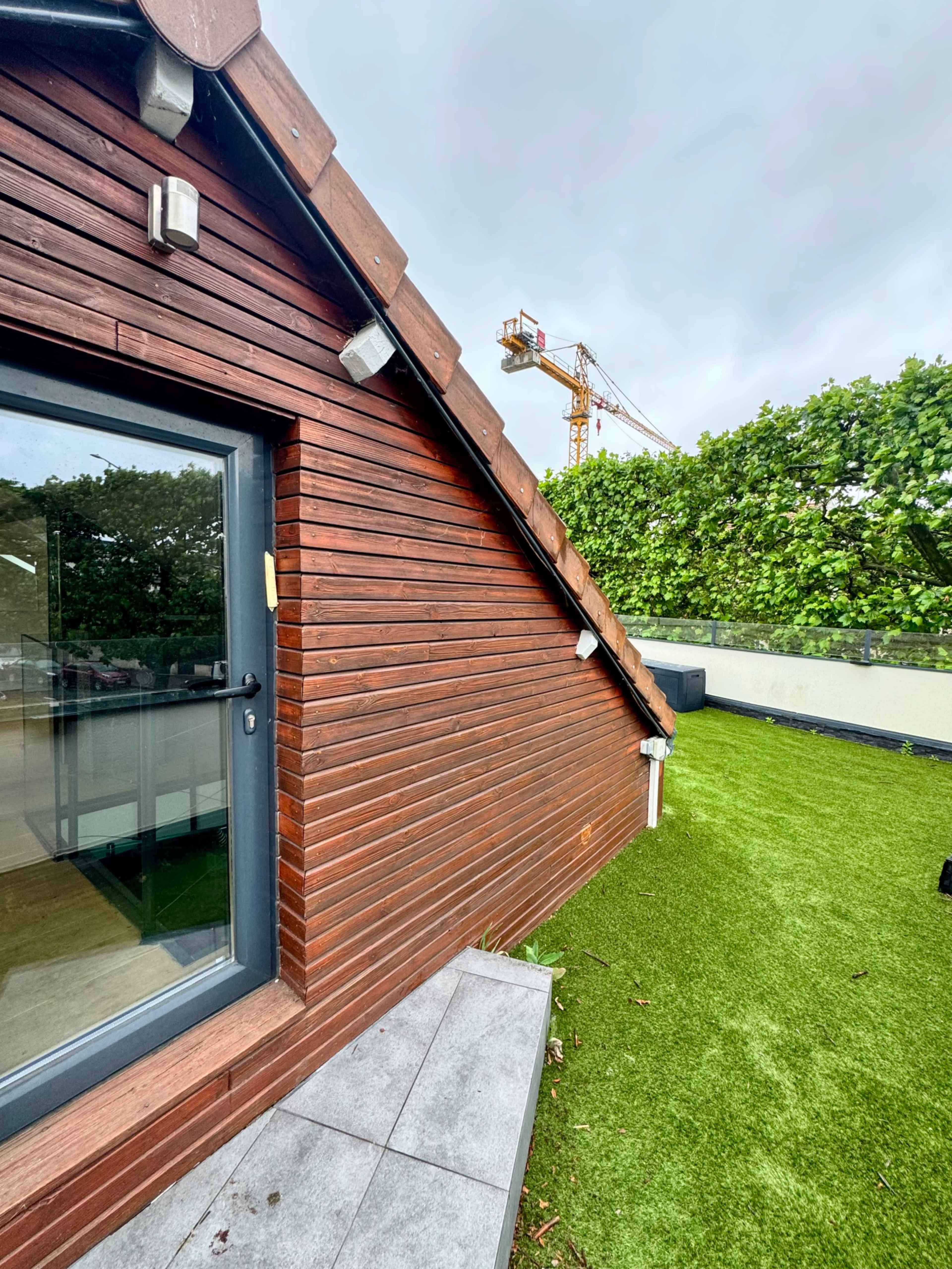 The image shows the side of a wooden building with a sloped roof, adjacent to a green lawn and a construction crane in the background.