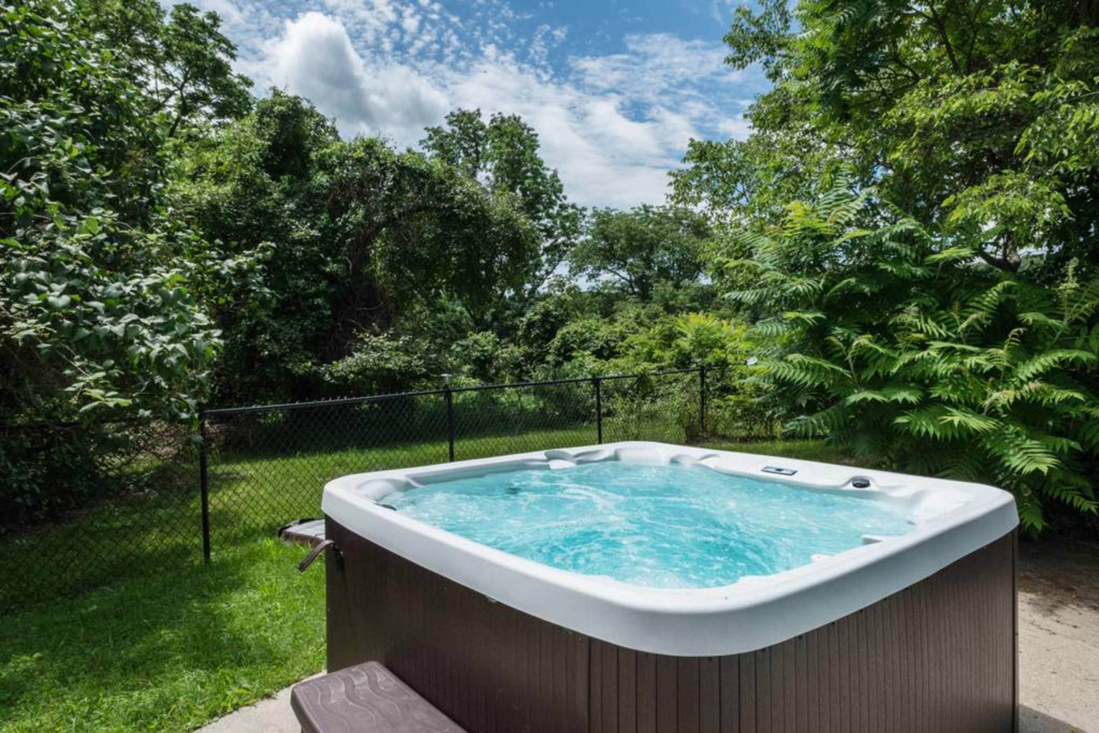 A hot tub filled with water is positioned on a patio in a backyard surrounded by trees and a fence.