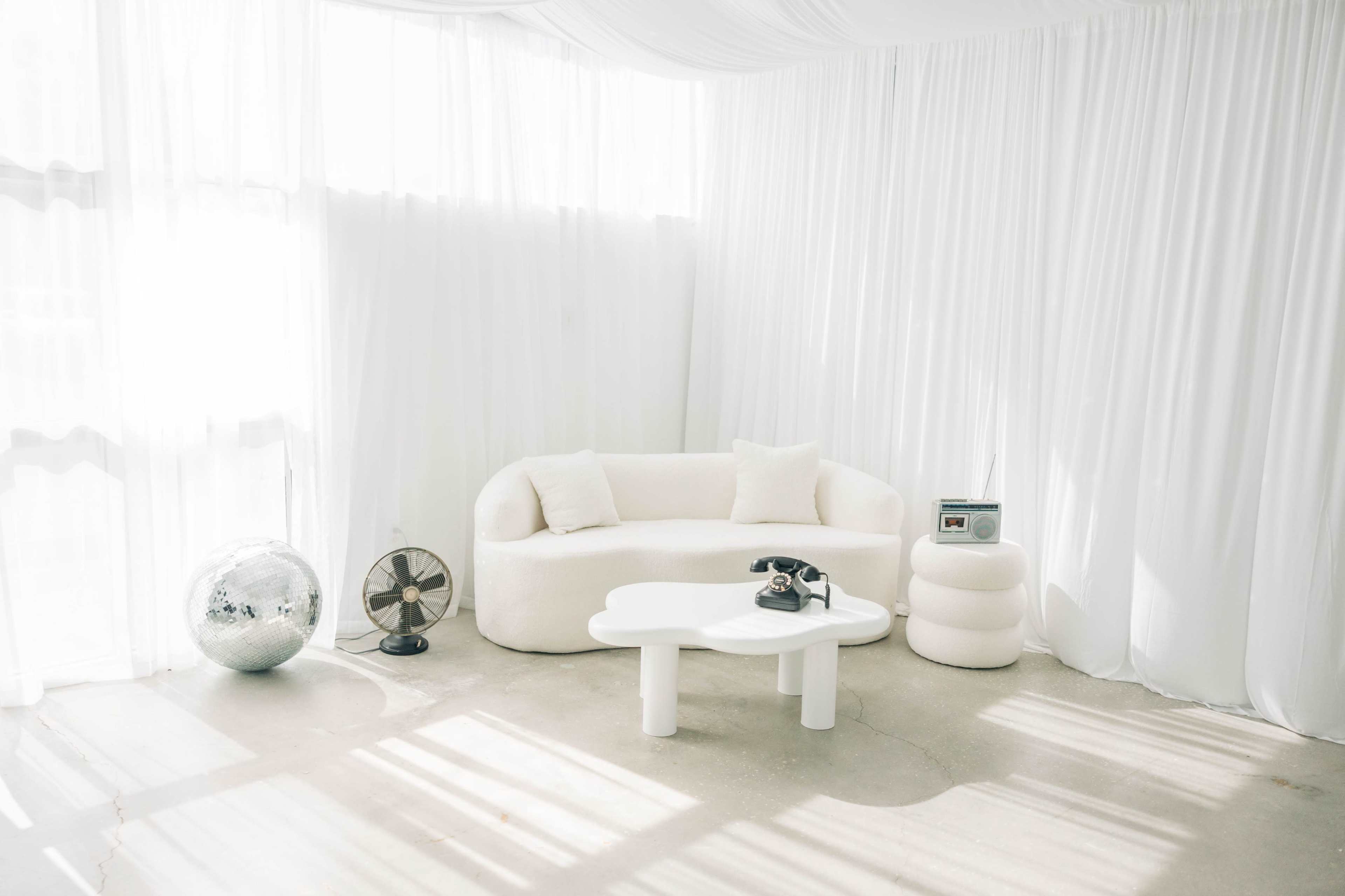 A minimalist living space features a white sofa, a round table, and a stool, all set against sheer white curtains and soft lighting.