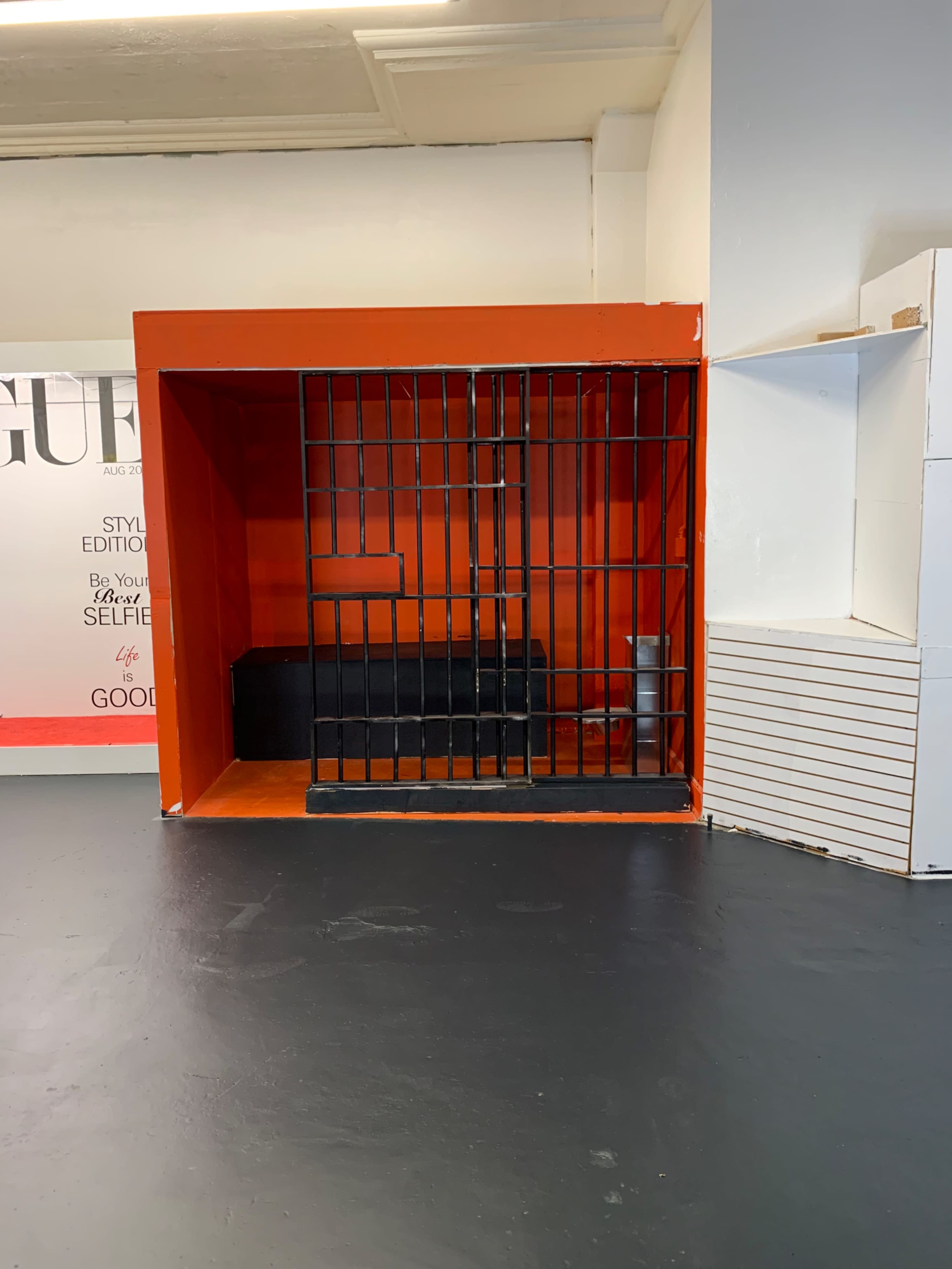 A small, empty cell with a black barred gate set against an orange background, located in a room with white walls and a black floor.