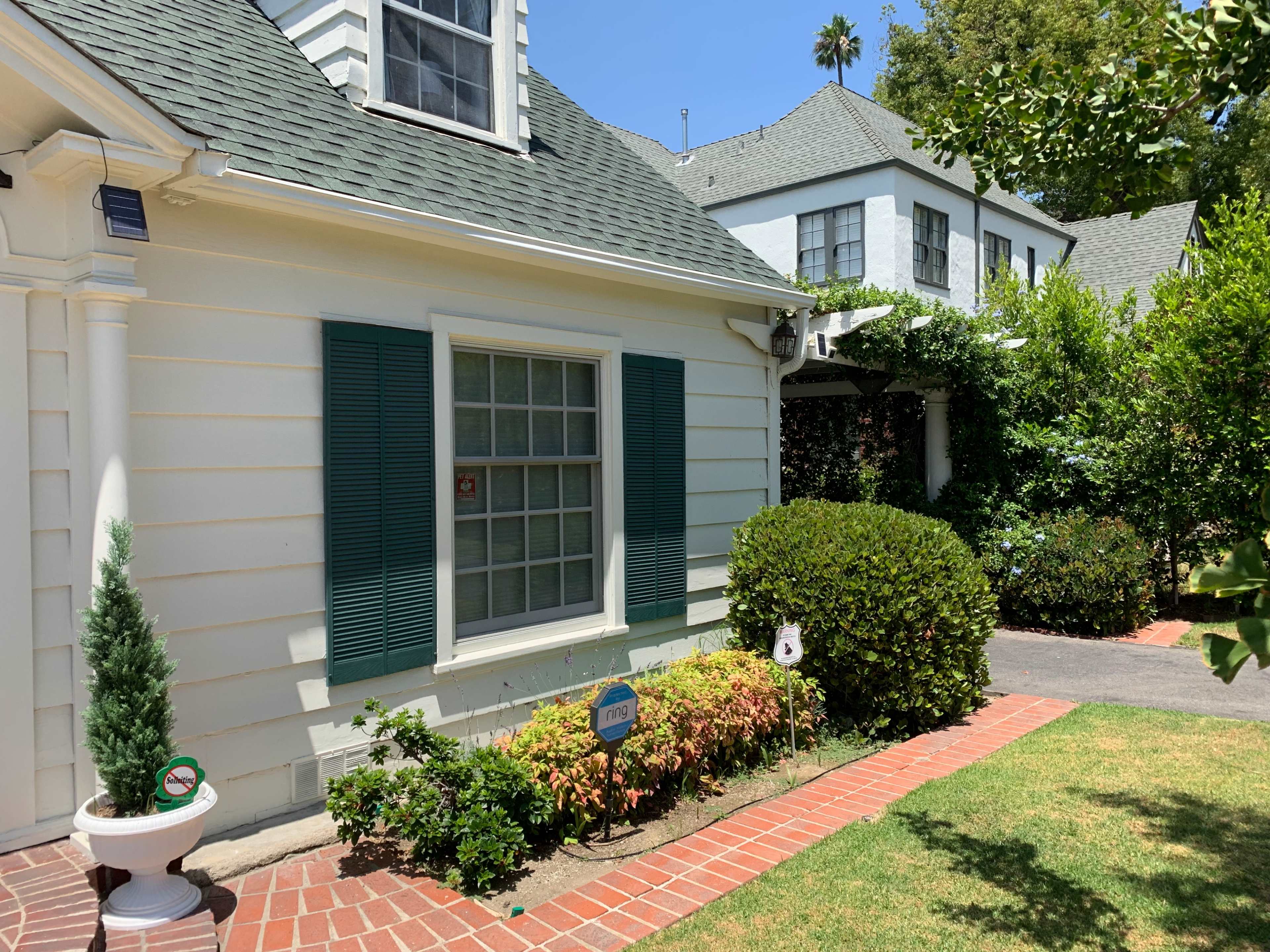 A white house with green shutters is surrounded by well-maintained landscaping and a brick walkway.