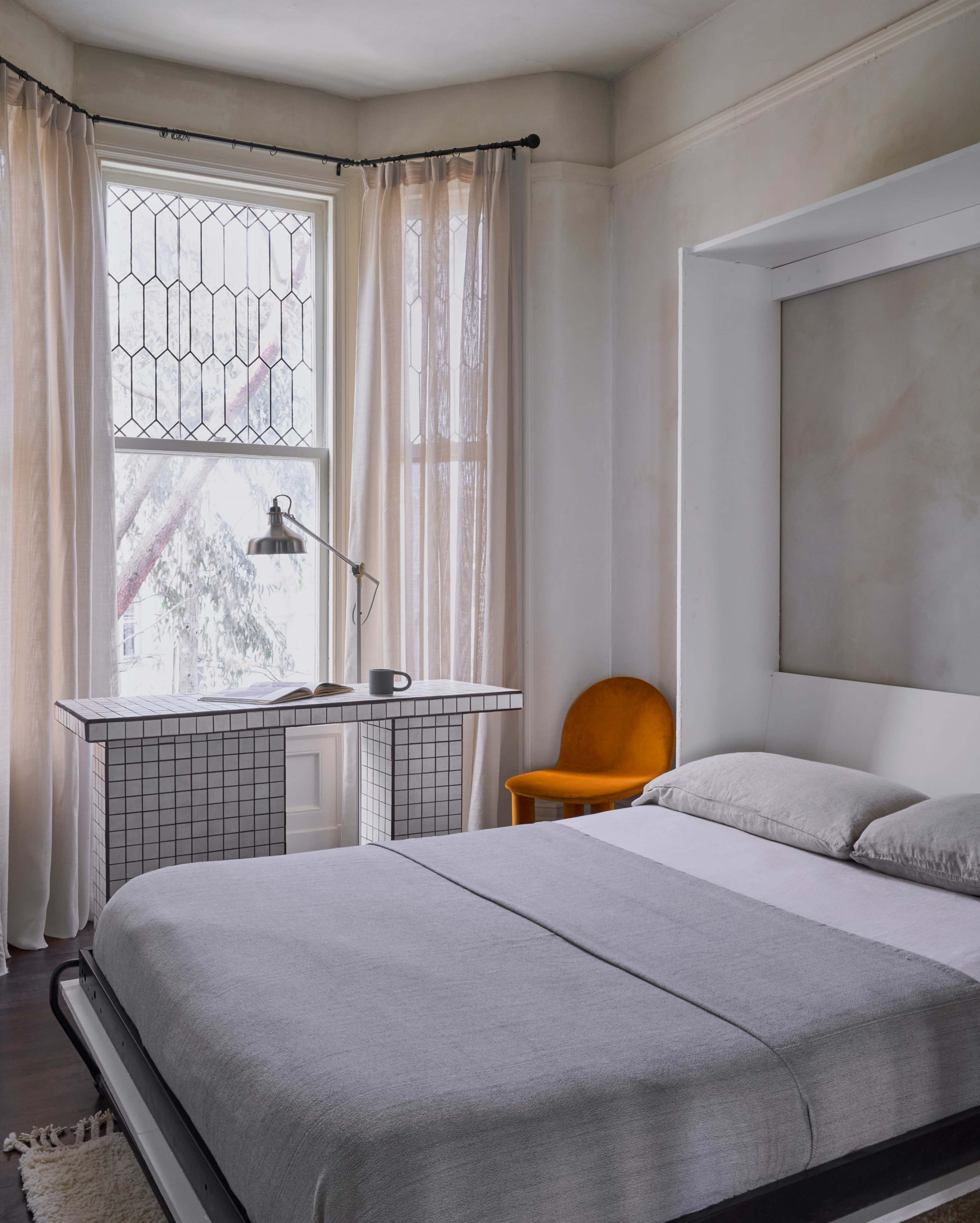 A minimalist bedroom features a bed with gray linens, a white-paneled wall, large curtained windows, and a desk with a grid-patterned surface beside an orange chair.
