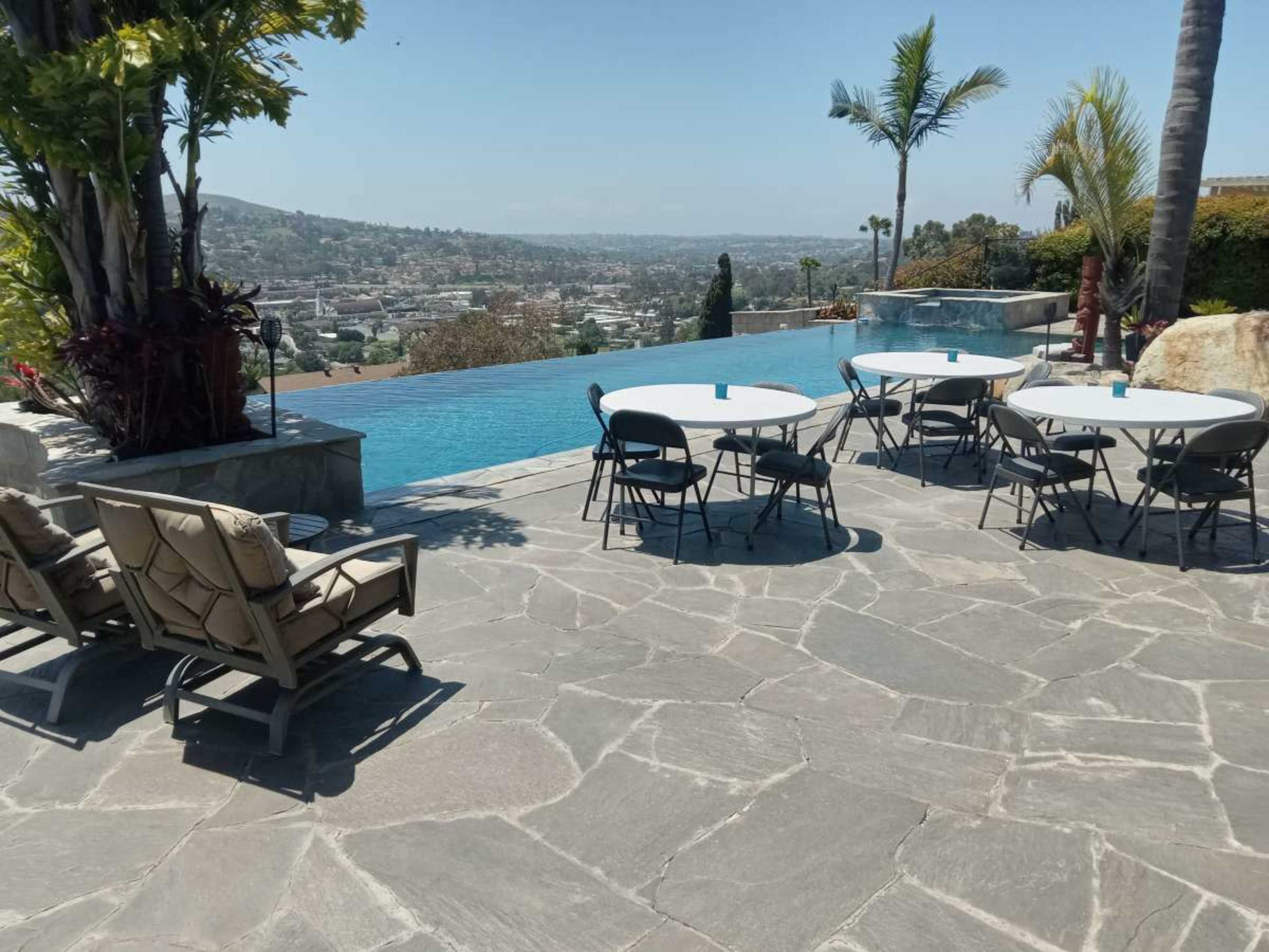 A patio area features several round tables and chairs beside a large infinity pool overlooking a cityscape.