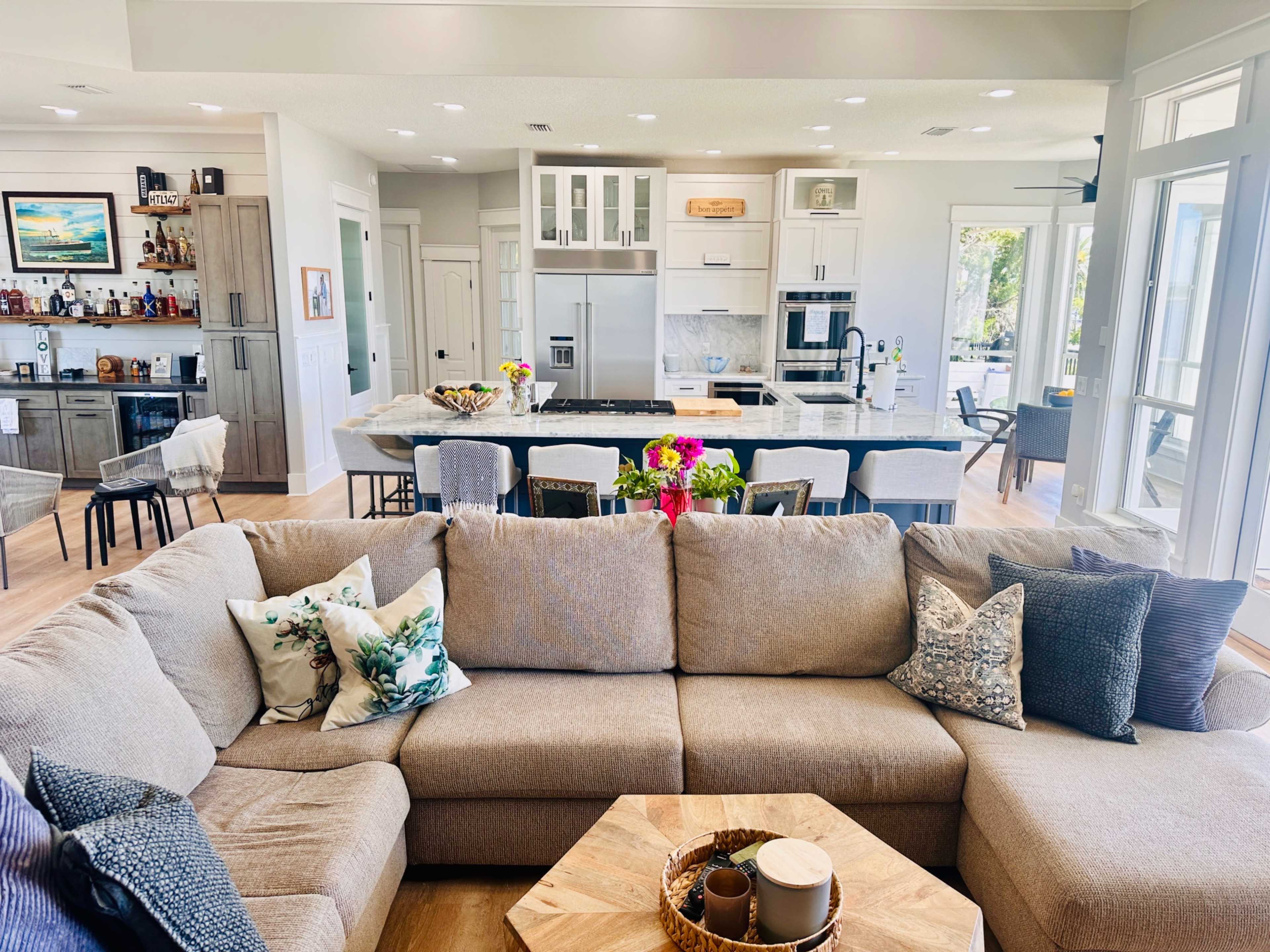 A large, modern living room features a beige sectional sofa, a wooden coffee table, and an open kitchen with a central island and bar seating in the background.