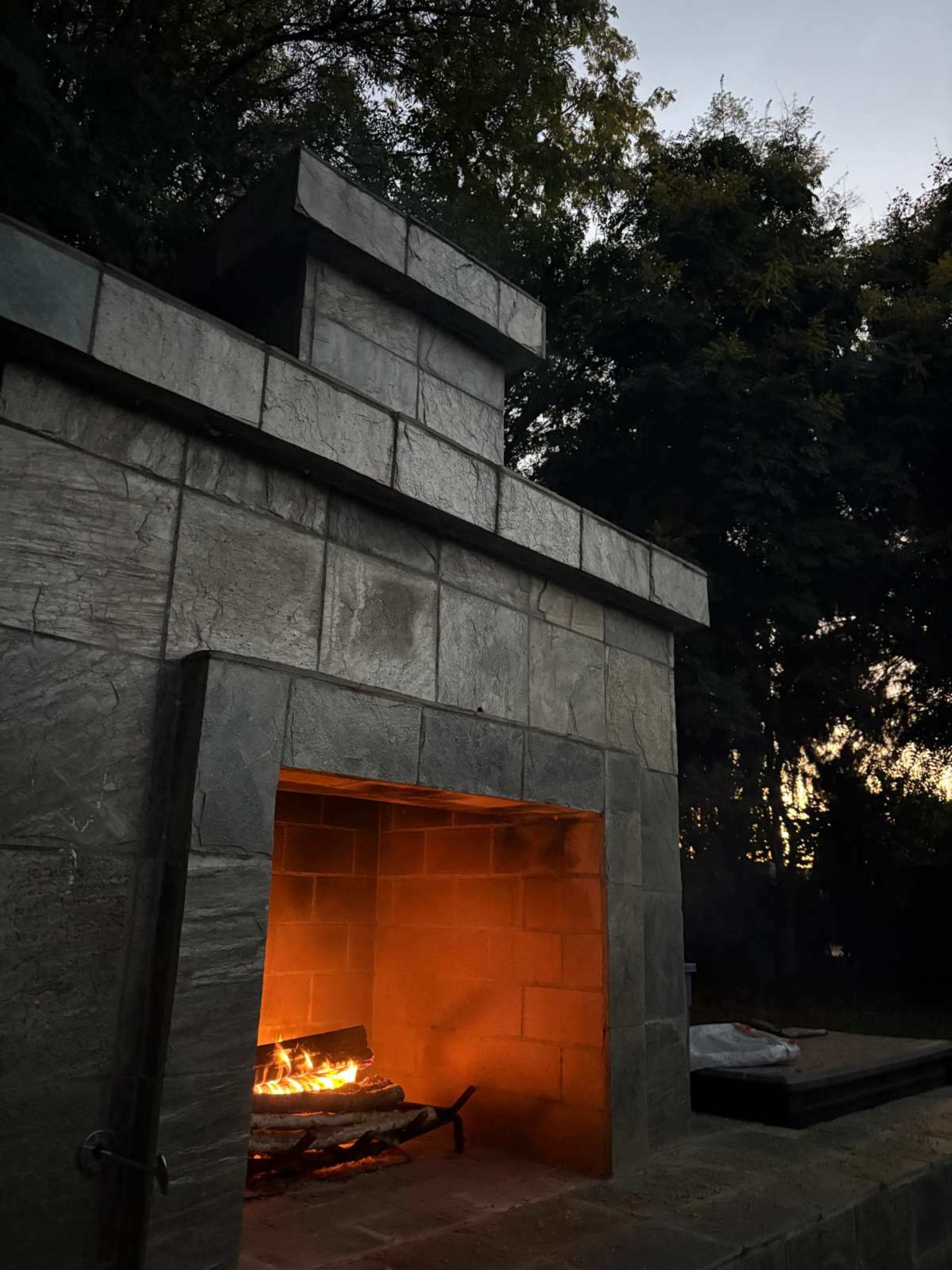 A stone outdoor fireplace with a visible fire casts a warm glow in the dusk.