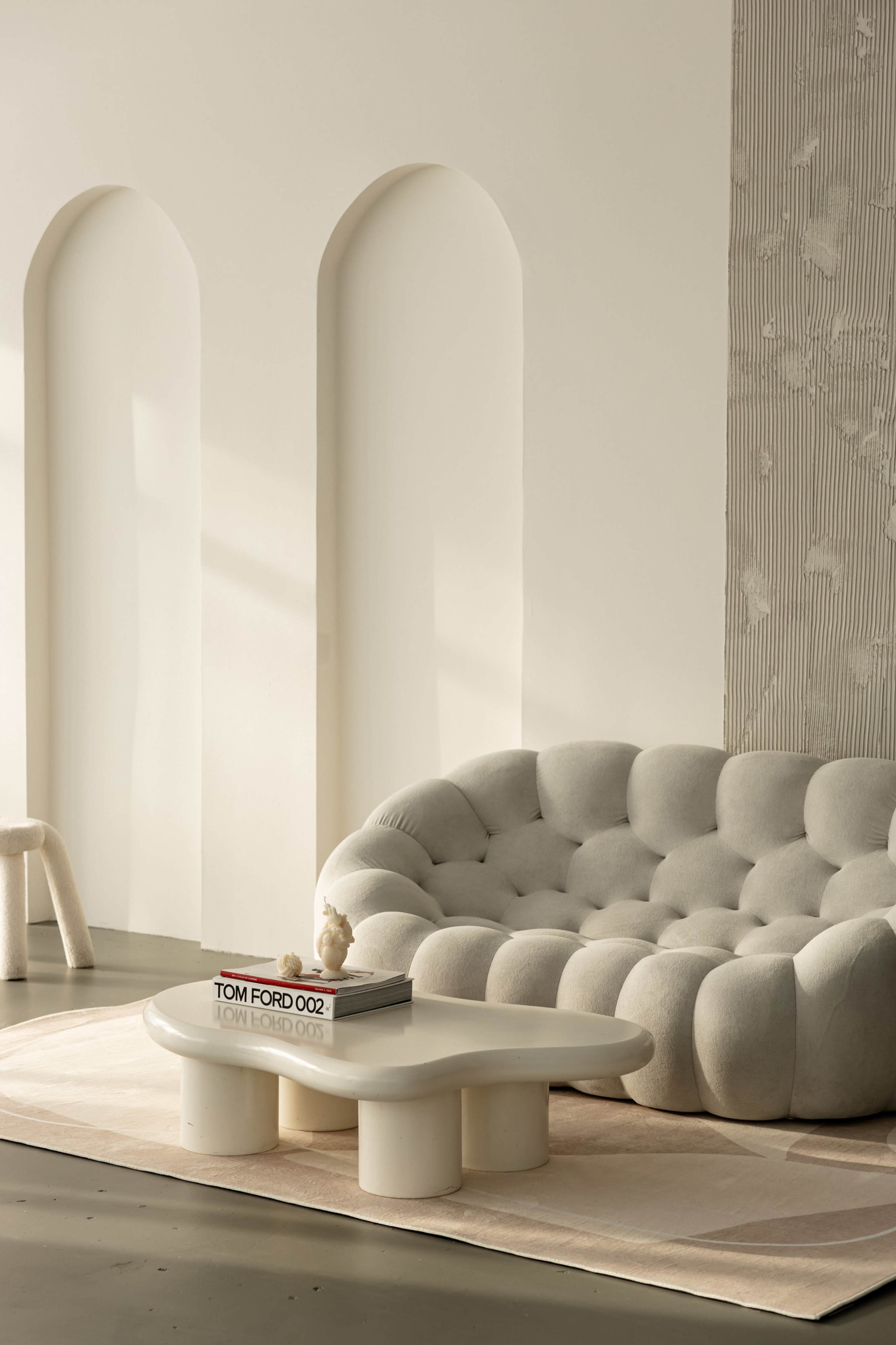 The image depicts a cozy living room featuring a plush, light-colored sofa, a round coffee table, and a soft area rug against a minimalist wall with archways.