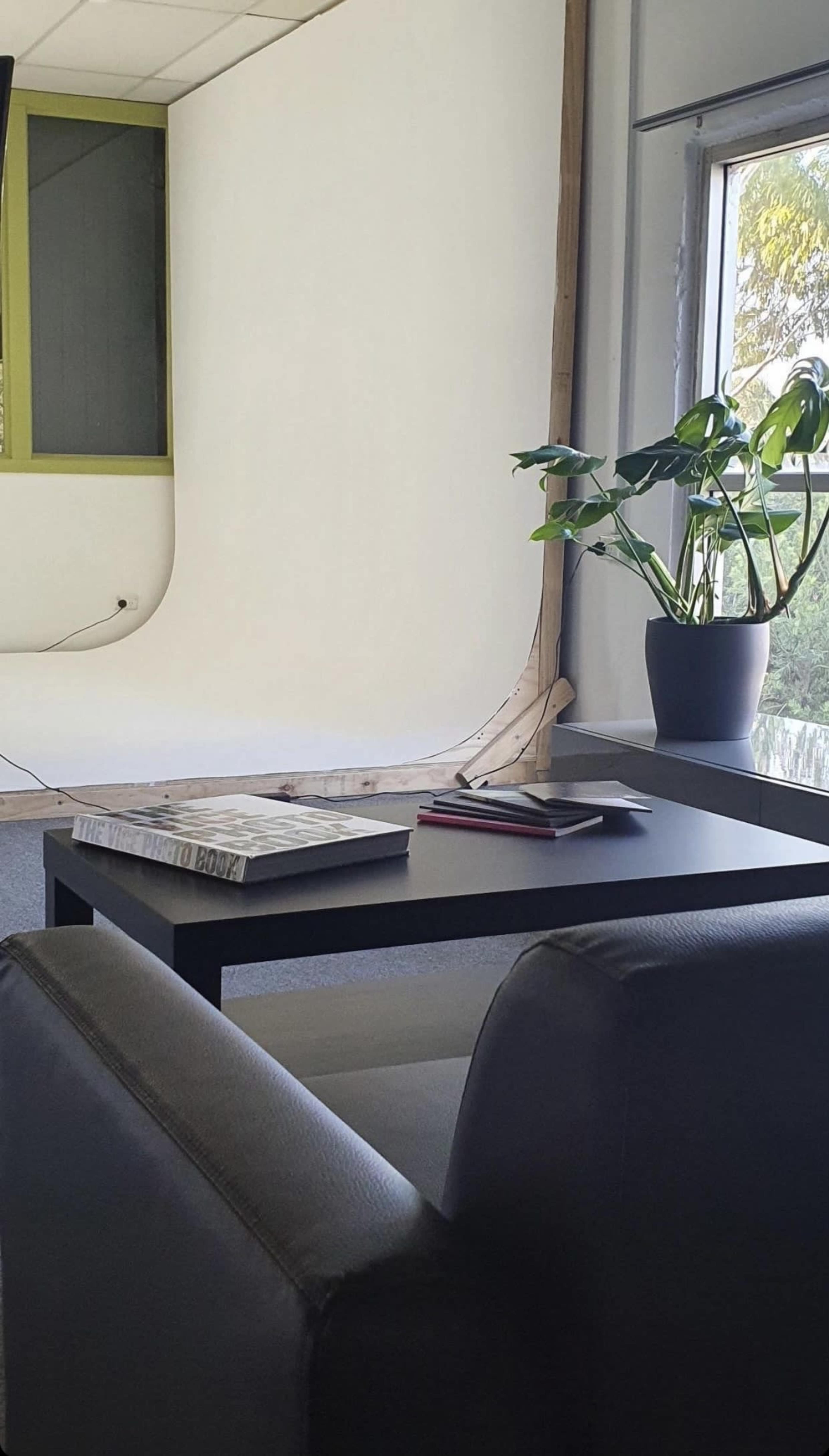 A modern photography studio setup with a seamless white backdrop, a black table holding magazines, and a potted plant near a large window.