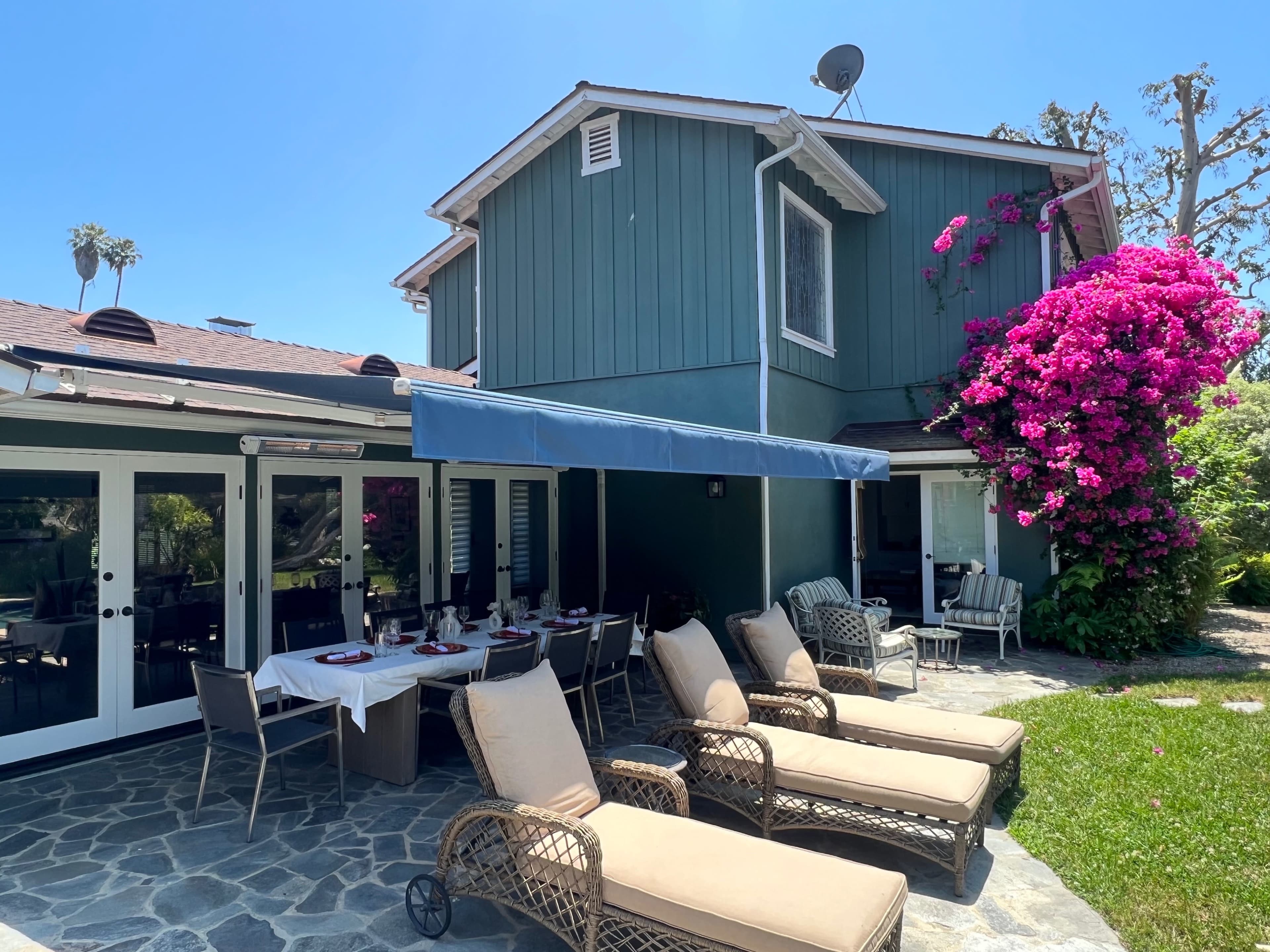 A patio with lounge chairs and a dining area is situated next to a green house adorned with blooming bougainvillea.