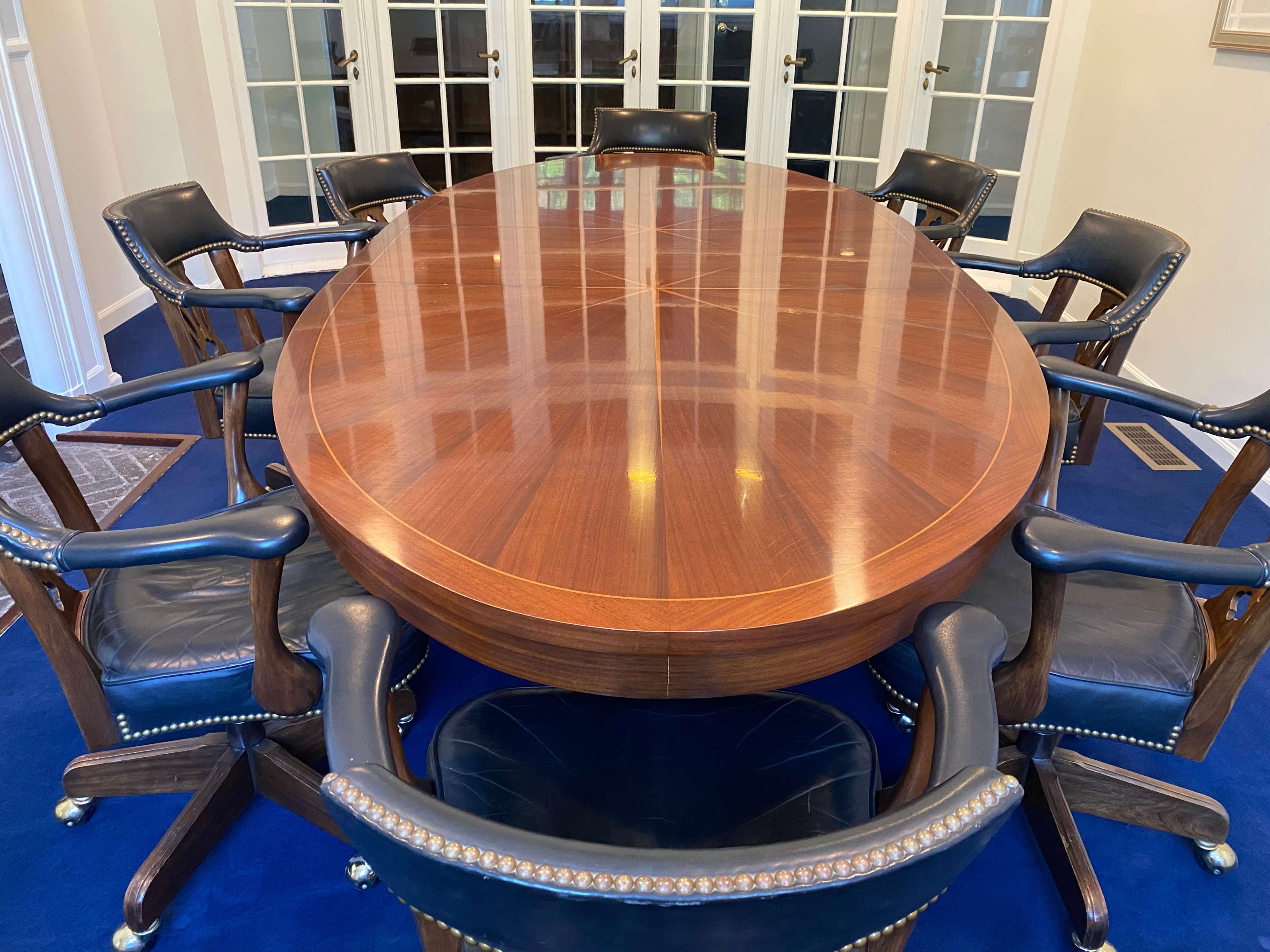 The image shows a large oval wooden conference table surrounded by eight chairs in a well-lit room with glass doors.