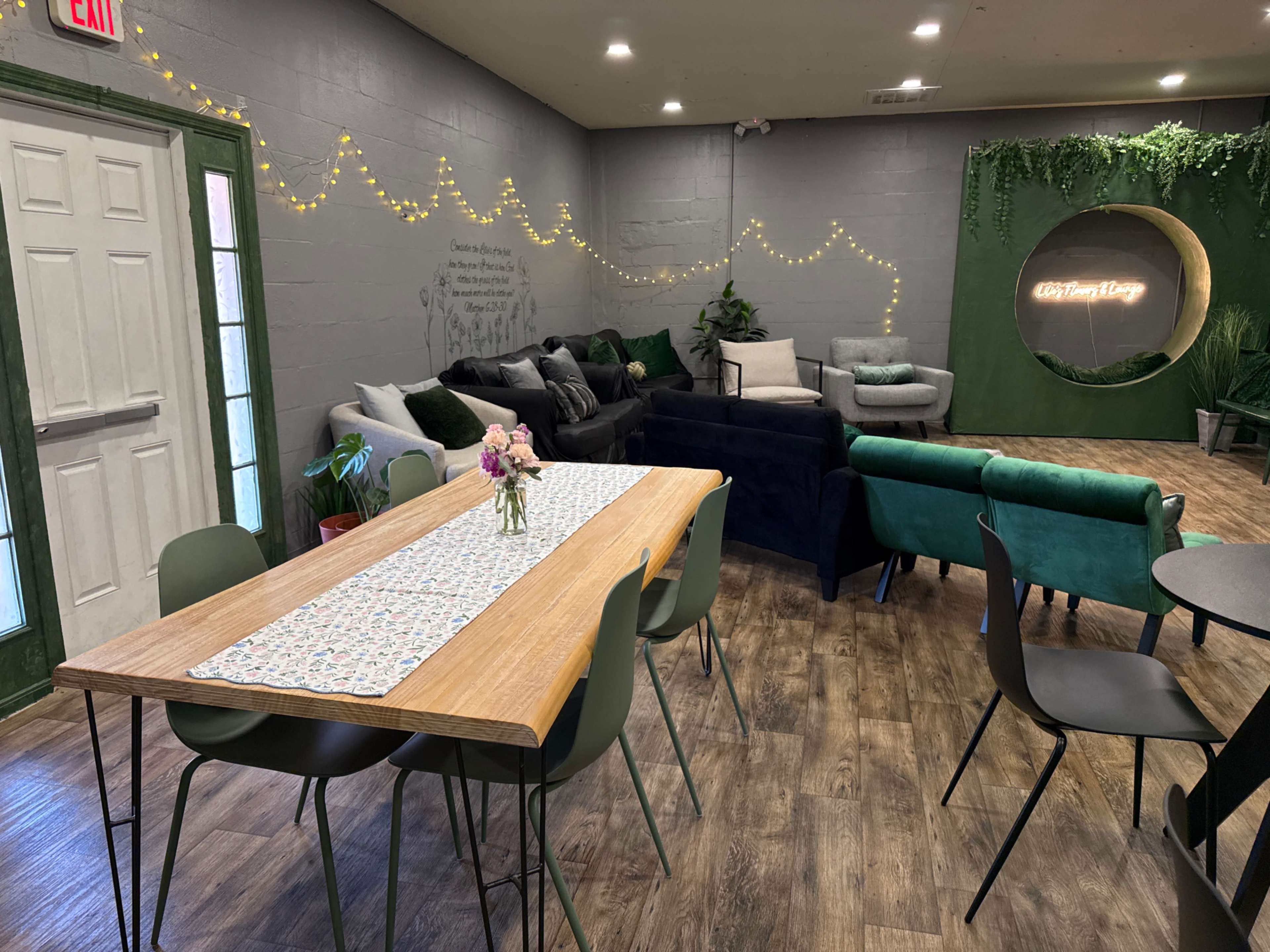 The image shows a modern interior space featuring a long wooden table surrounded by green chairs, with a cozy seating area containing dark sofas and decorative plants.