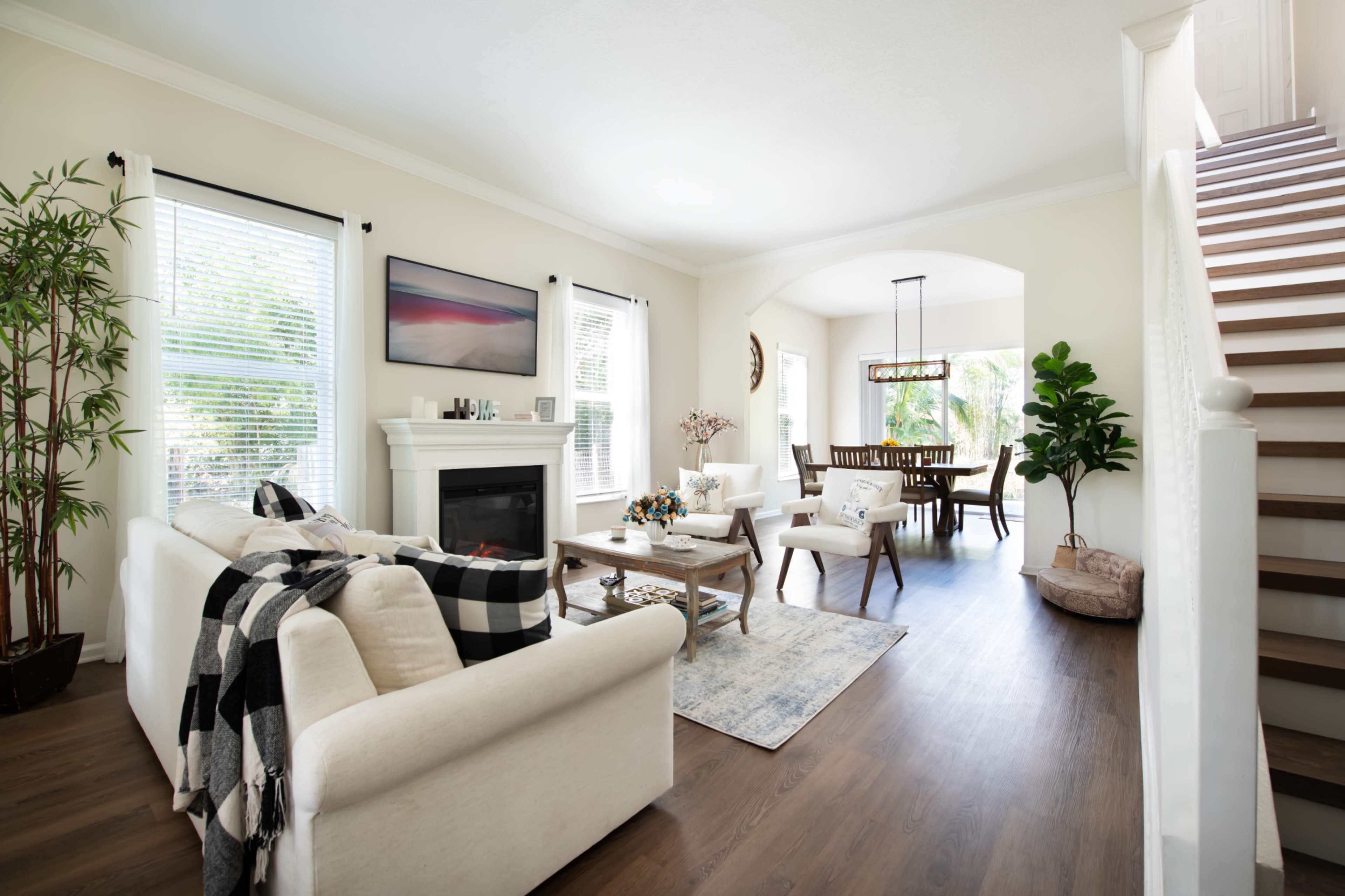 A cozy living room features a white sofa, a coffee table, a fireplace, and a dining area with a large table, all set in a bright, open space with a staircase leading to the upper floor.