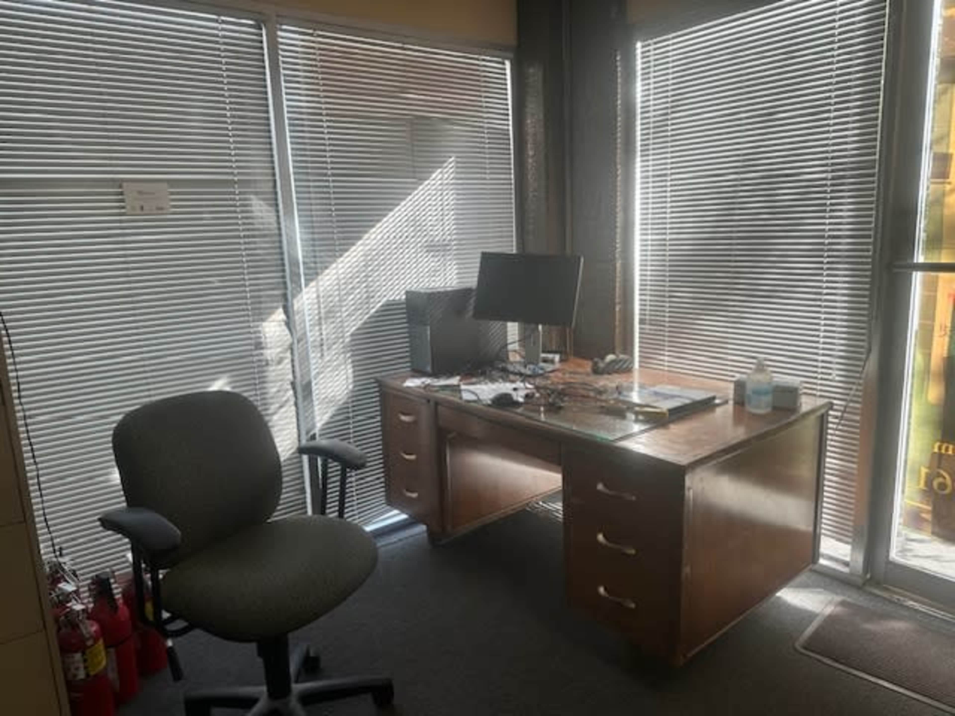 The image shows an office space featuring a wooden desk with a computer monitor, a swivel chair, and windows with blinds partially drawn.