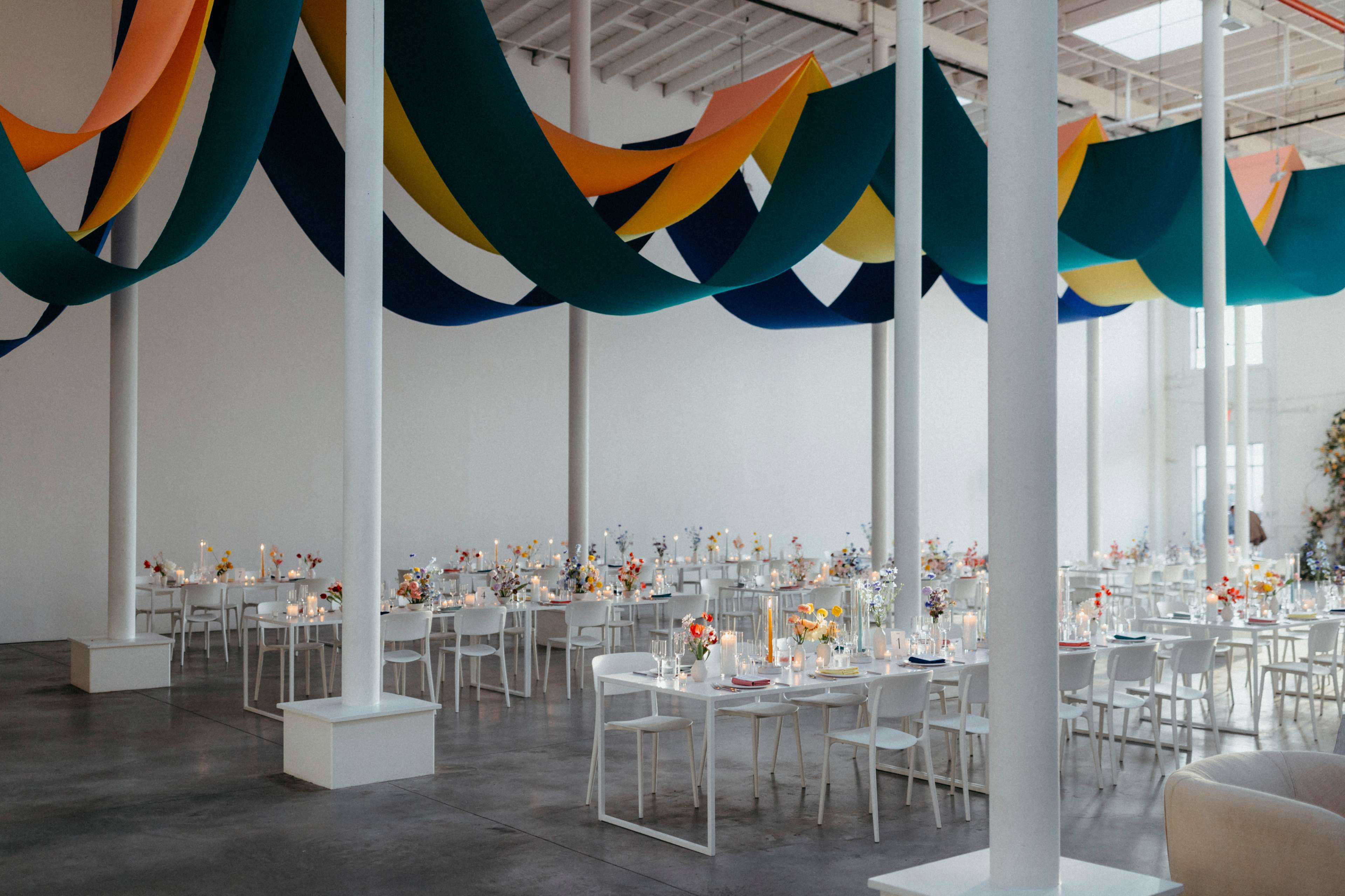 The spacious venue features tables adorned with flowers and candles, accompanied by colorful drapery hanging from the ceiling.