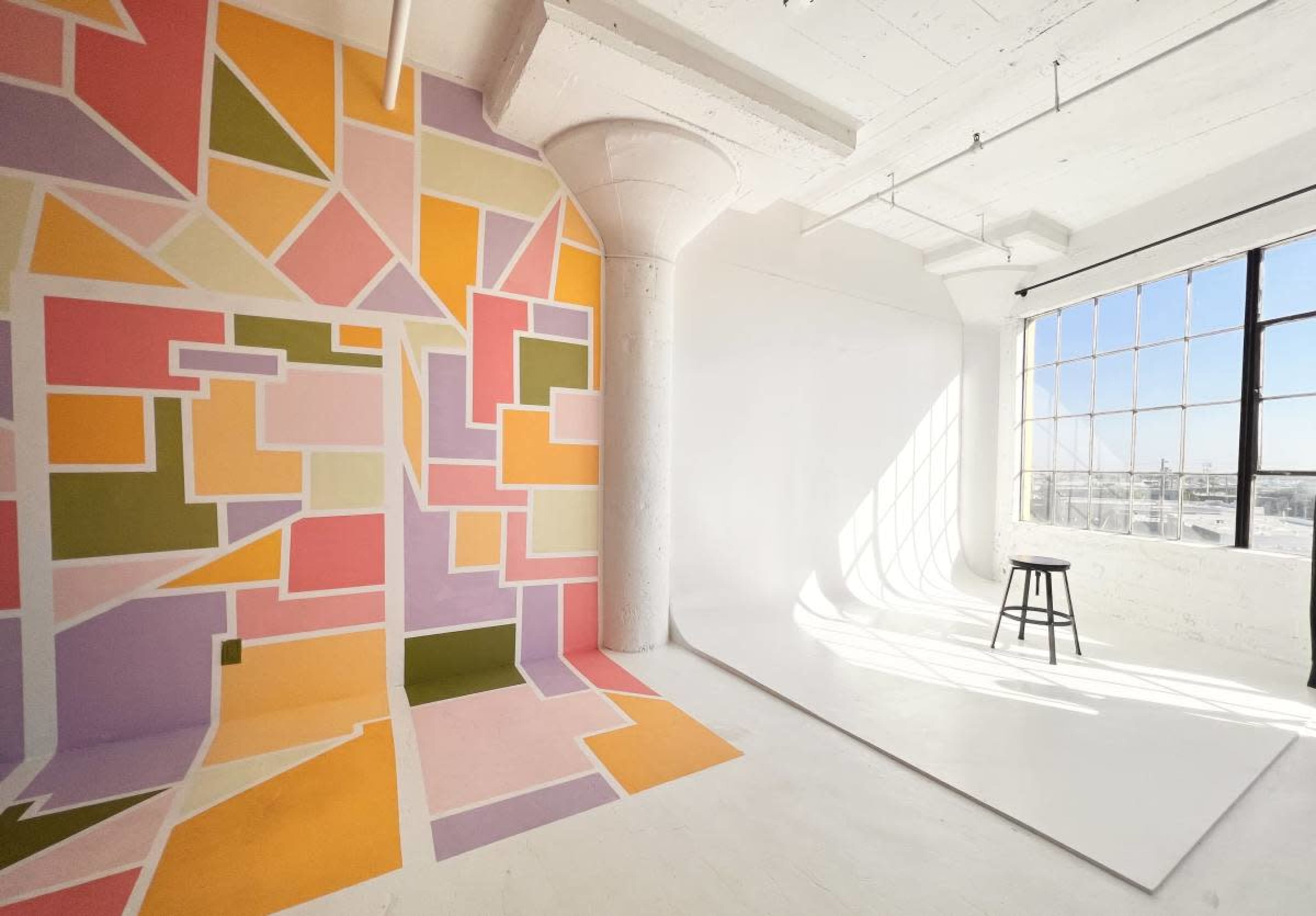 An artistically painted wall featuring geometric shapes in various colors alongside a bright, empty studio space with a stool and large windows.