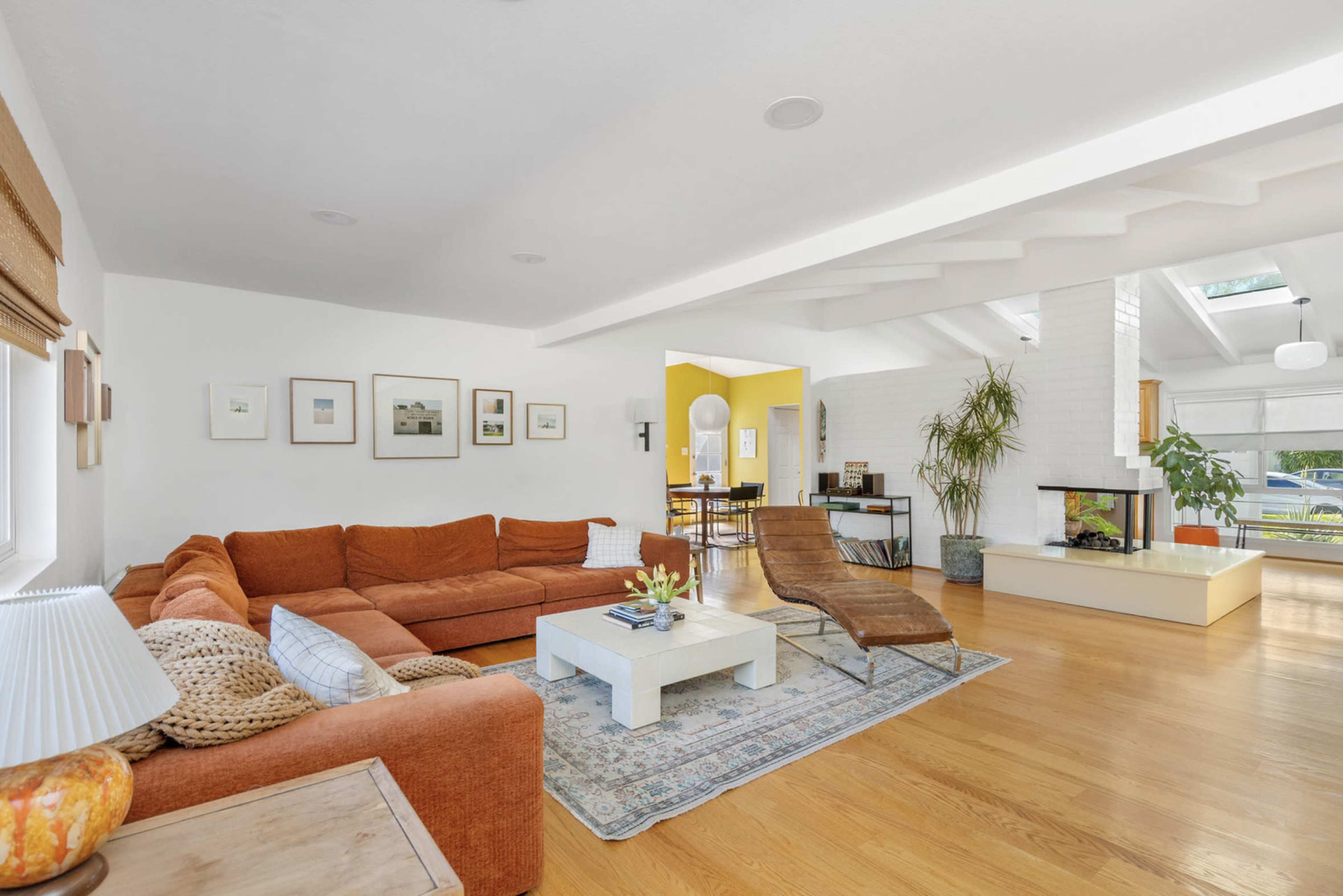 The image shows a spacious living room with a large orange sectional sofa, a glass-top coffee table, and a modern lounge chair, along with wood flooring and white walls.