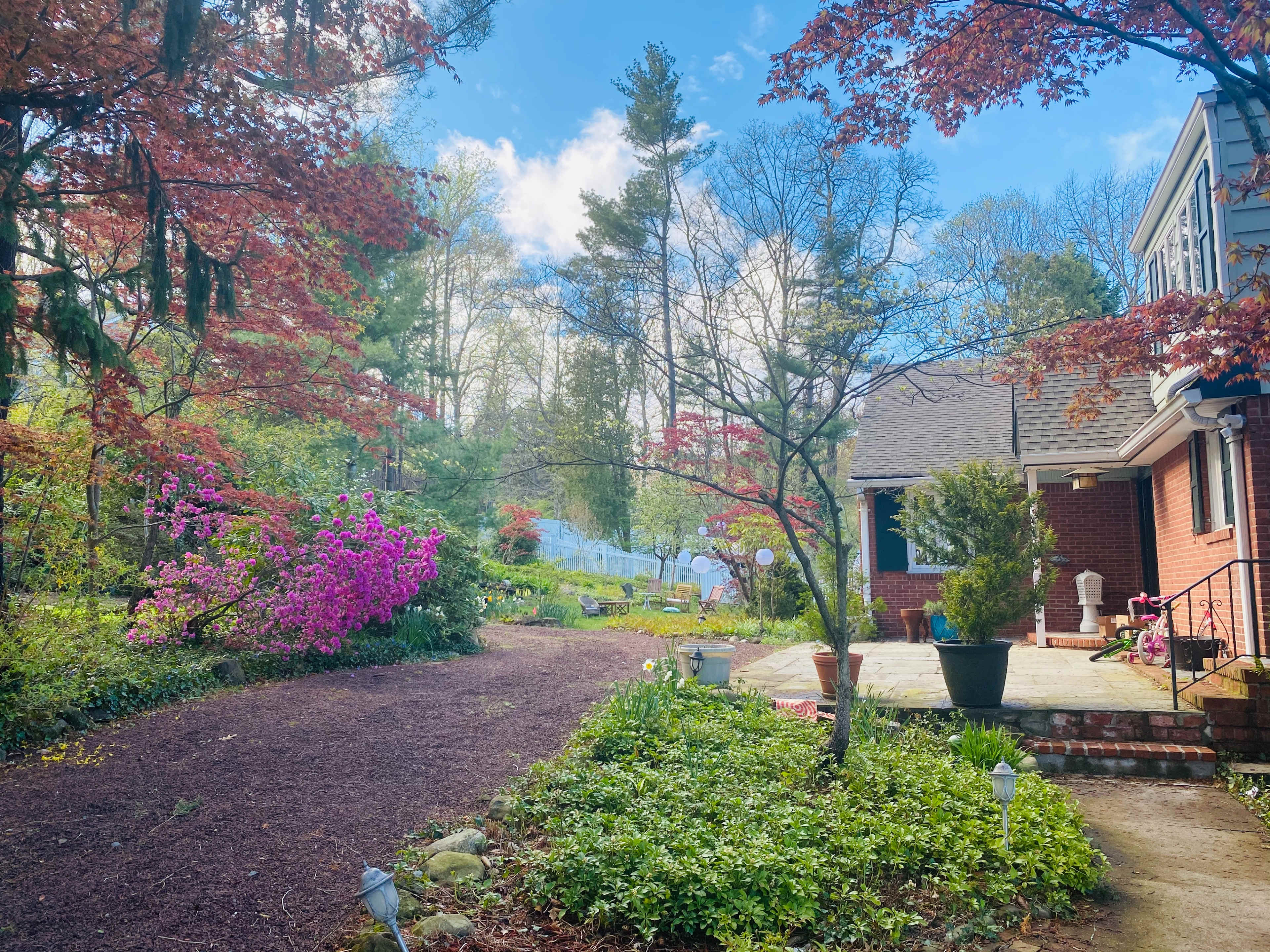 A winding gravel path leads through colorful blooming plants to a brick house surrounded by trees.