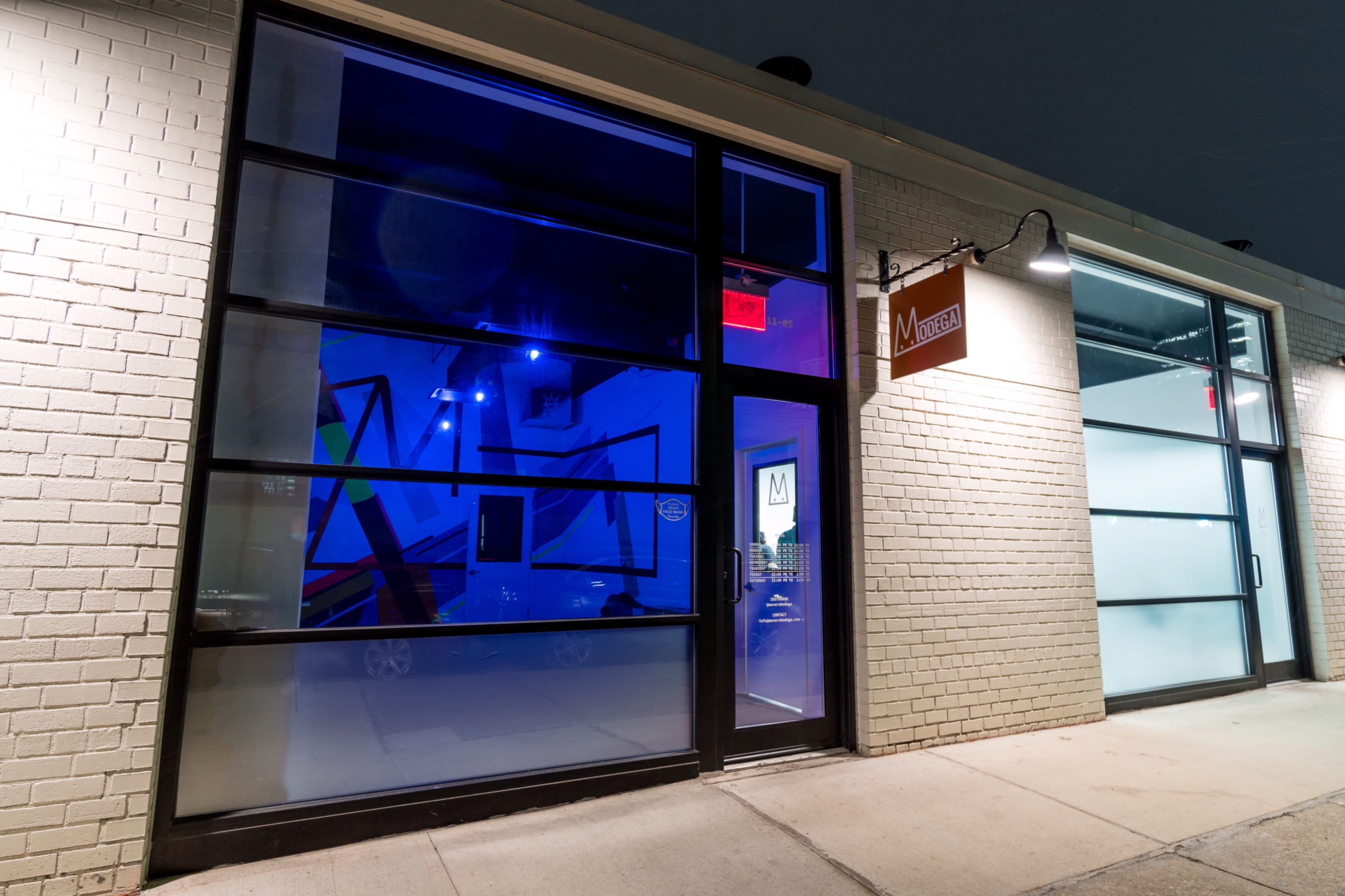 A modern storefront with large glass windows illuminated by blue lighting, featuring a sign that reads "M" above the entrance.
