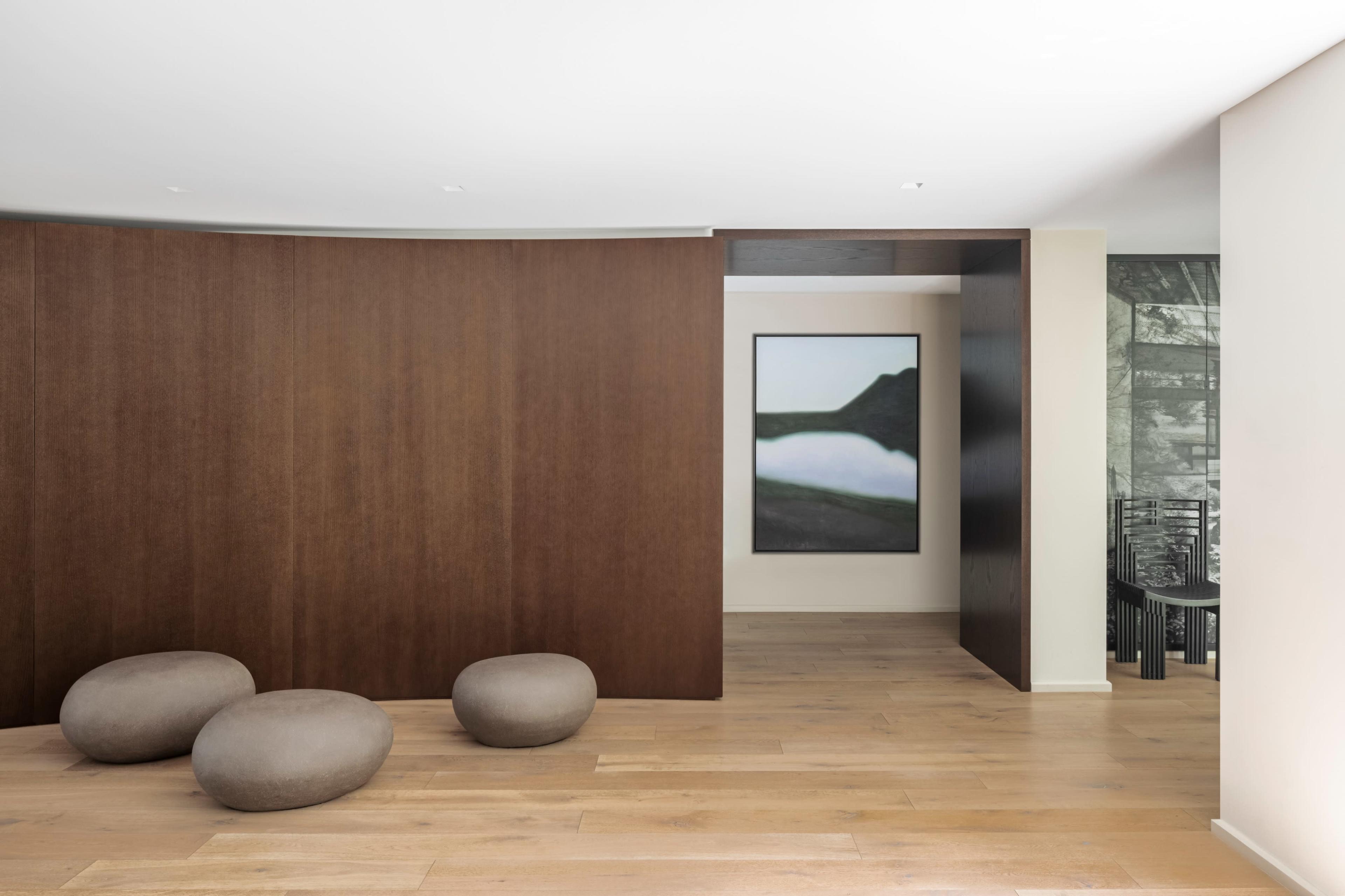 The image shows a modern interior space featuring smooth wooden flooring, rounded stone-like seating, and a large artwork on a wall in the background.
