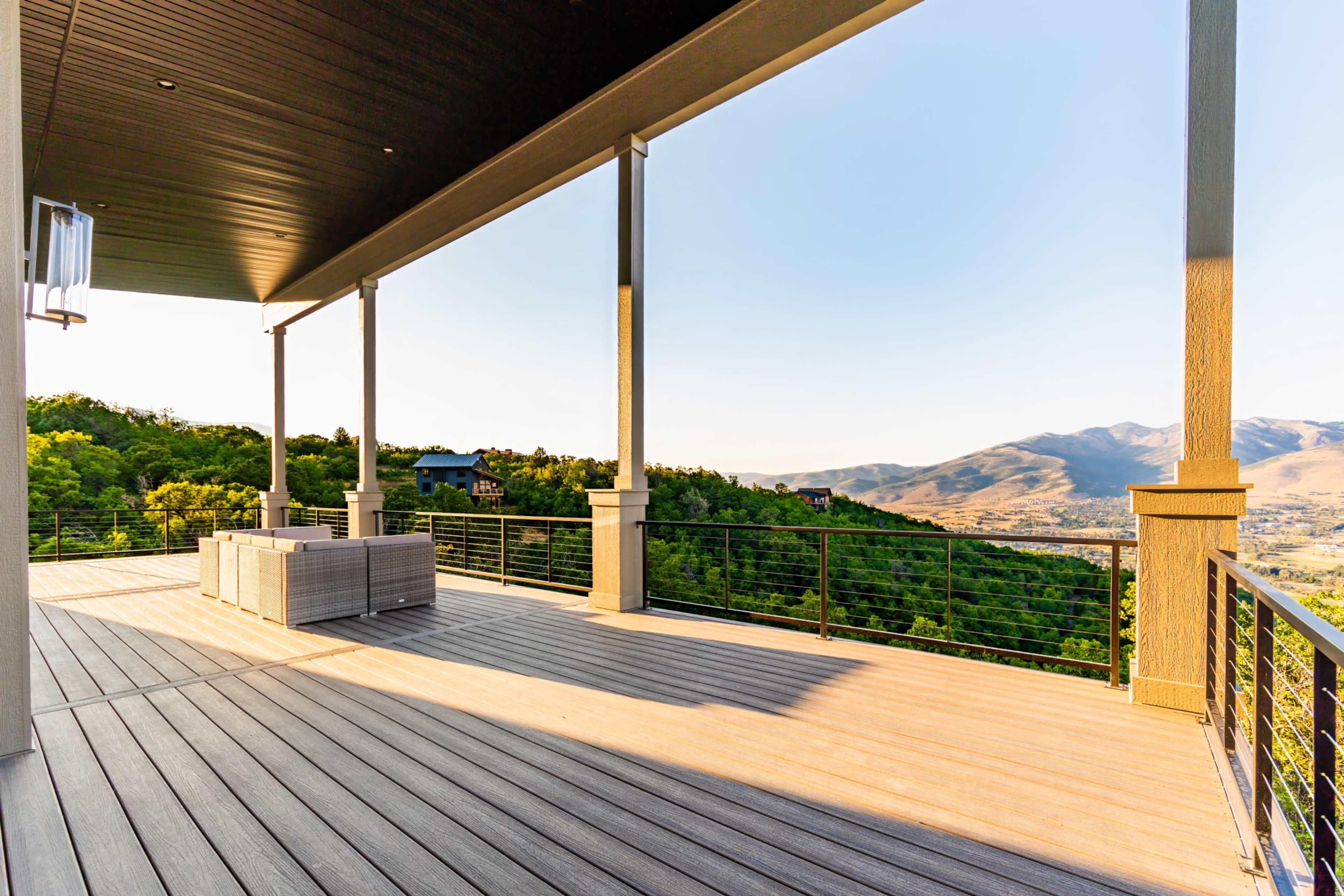 The image shows a spacious wooden deck with modern outdoor furniture, overlooking a lush green hillside and distant mountain range under a clear blue sky.